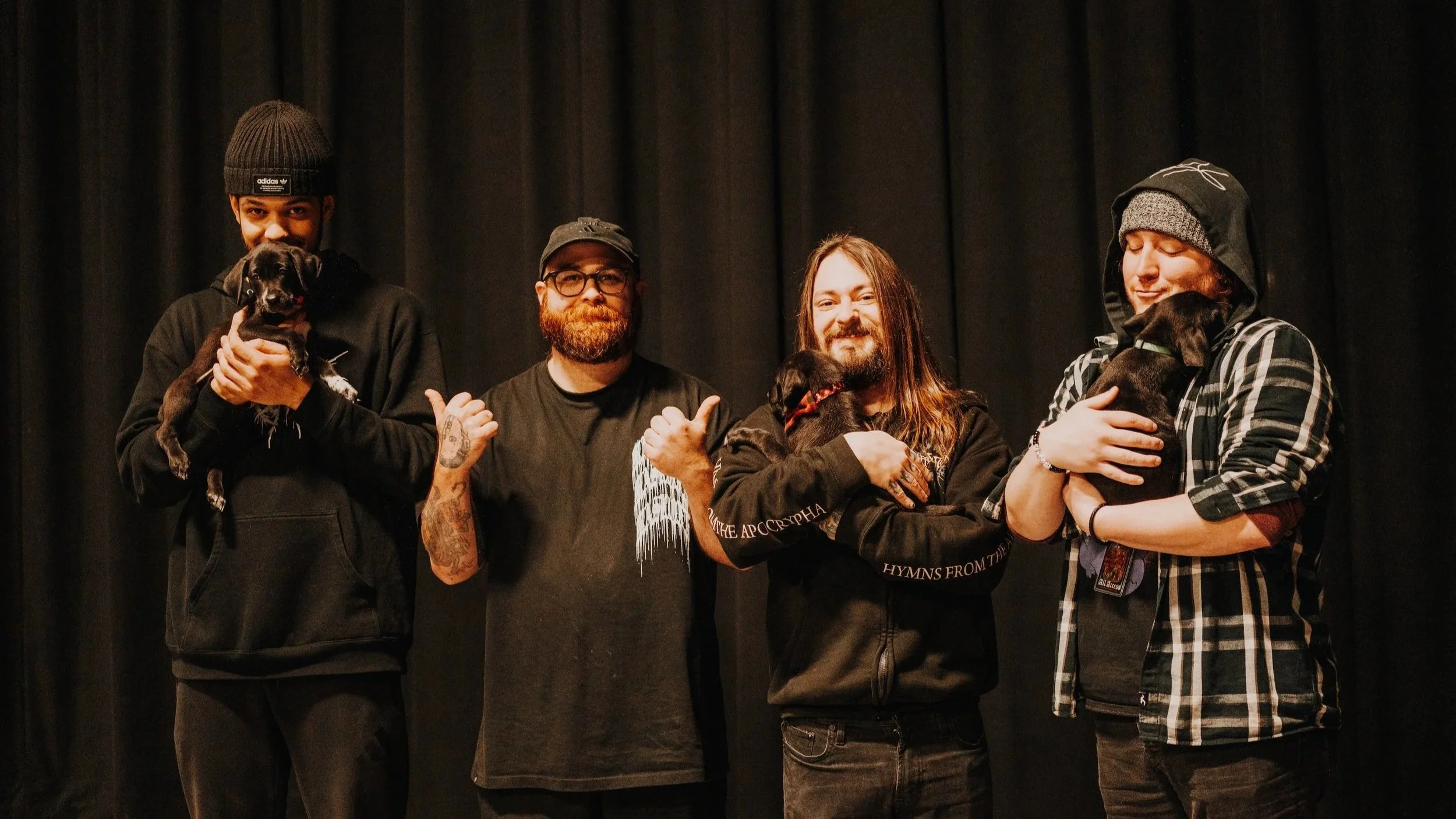 Four men standing in front of a black curtain, each holding a small black and brown dog. The men are smiling, making gestures, and wearing casual dark clothing and beanies or hats.