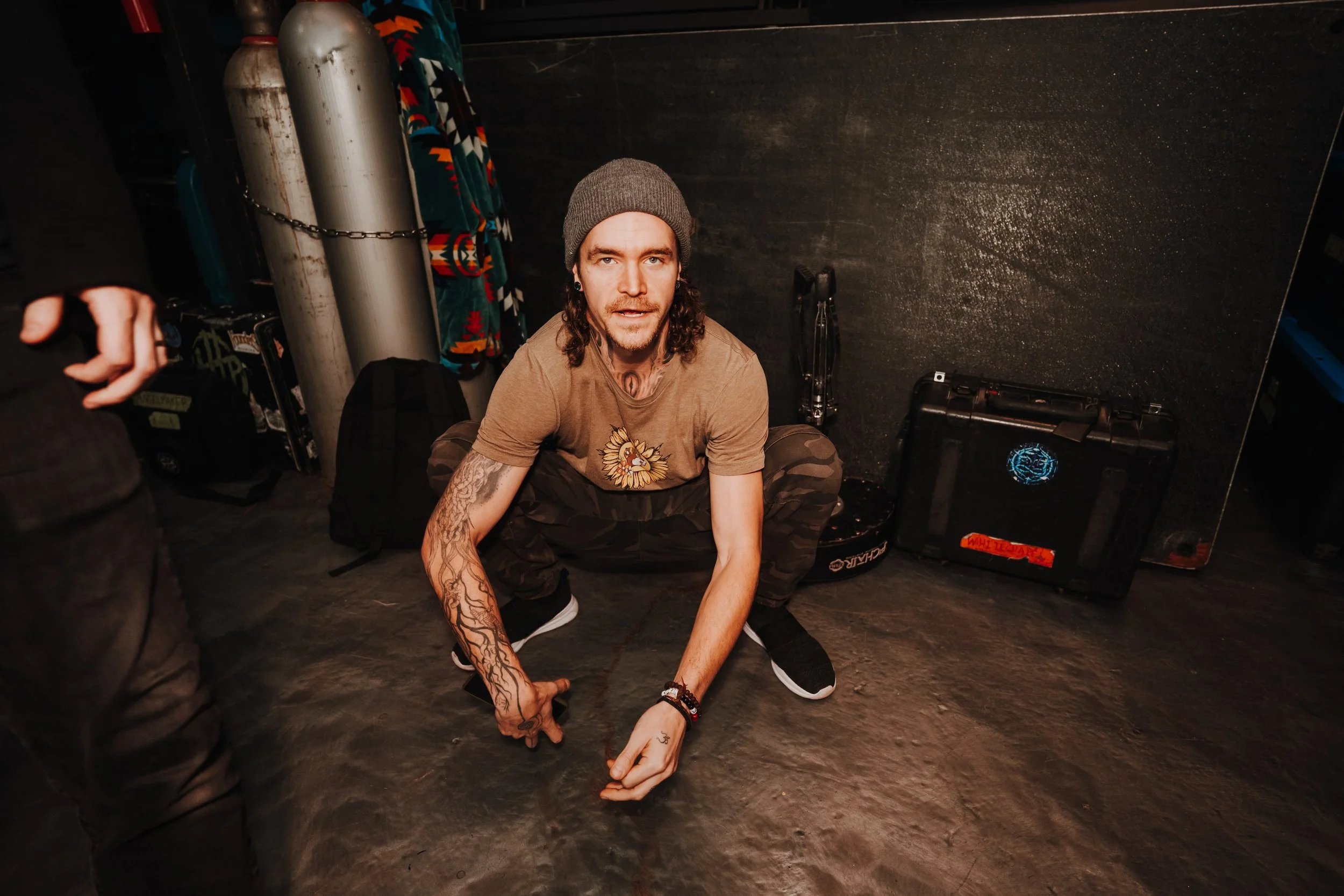 A man with long curly hair wearing a gray beanie, a tan T-shirt with a lion graphic, and camo pants is squatting on the floor, looking at the camera. He has tattoos on his right arm and is wearing bracelets. The background shows two large oxygen tanks, a black wall, and a closed case with a glowing blue sticker.