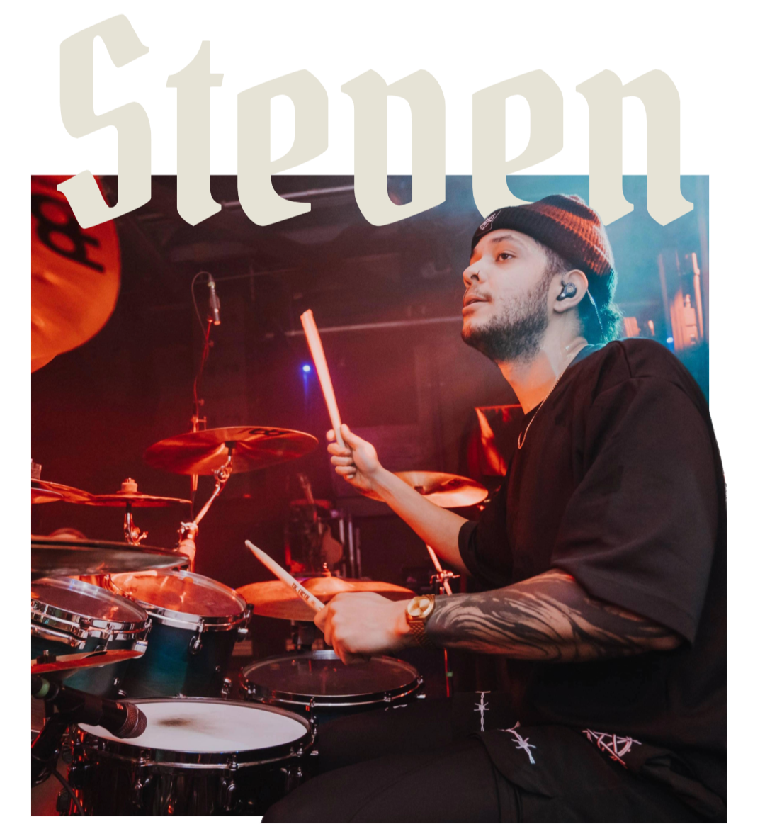A young man playing drums on stage, wearing a black beanie, black shirt, and a watch, with stage lighting in the background.
