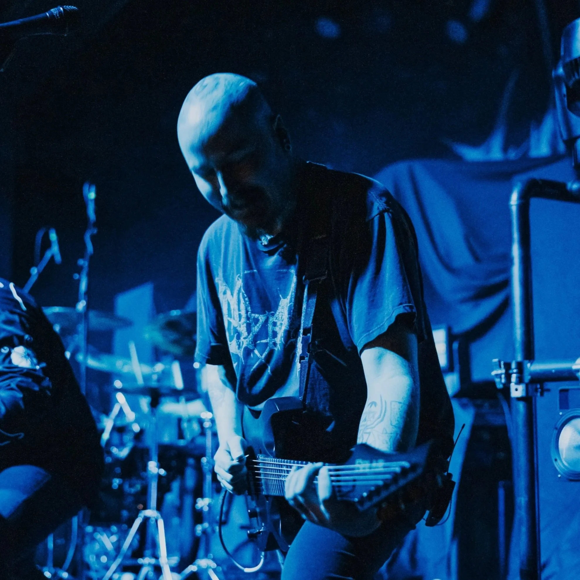 A person with a shaved head playing an electric guitar on stage, illuminated in blue light.