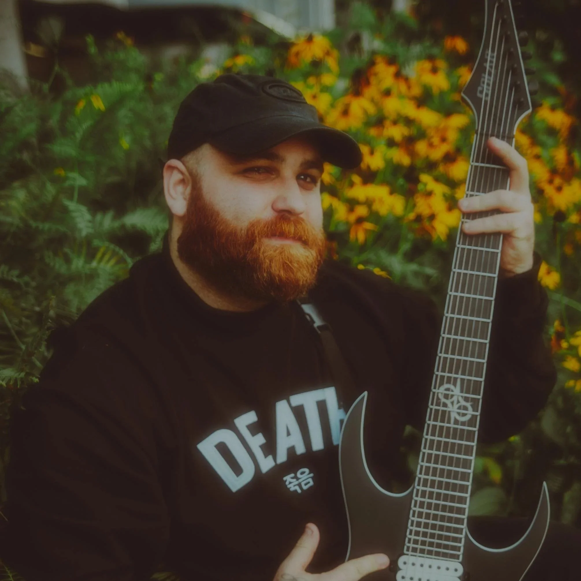 Man with a red beard wearing a black cap and shirt, holding a black electric guitar outdoors with yellow flowers in the background.