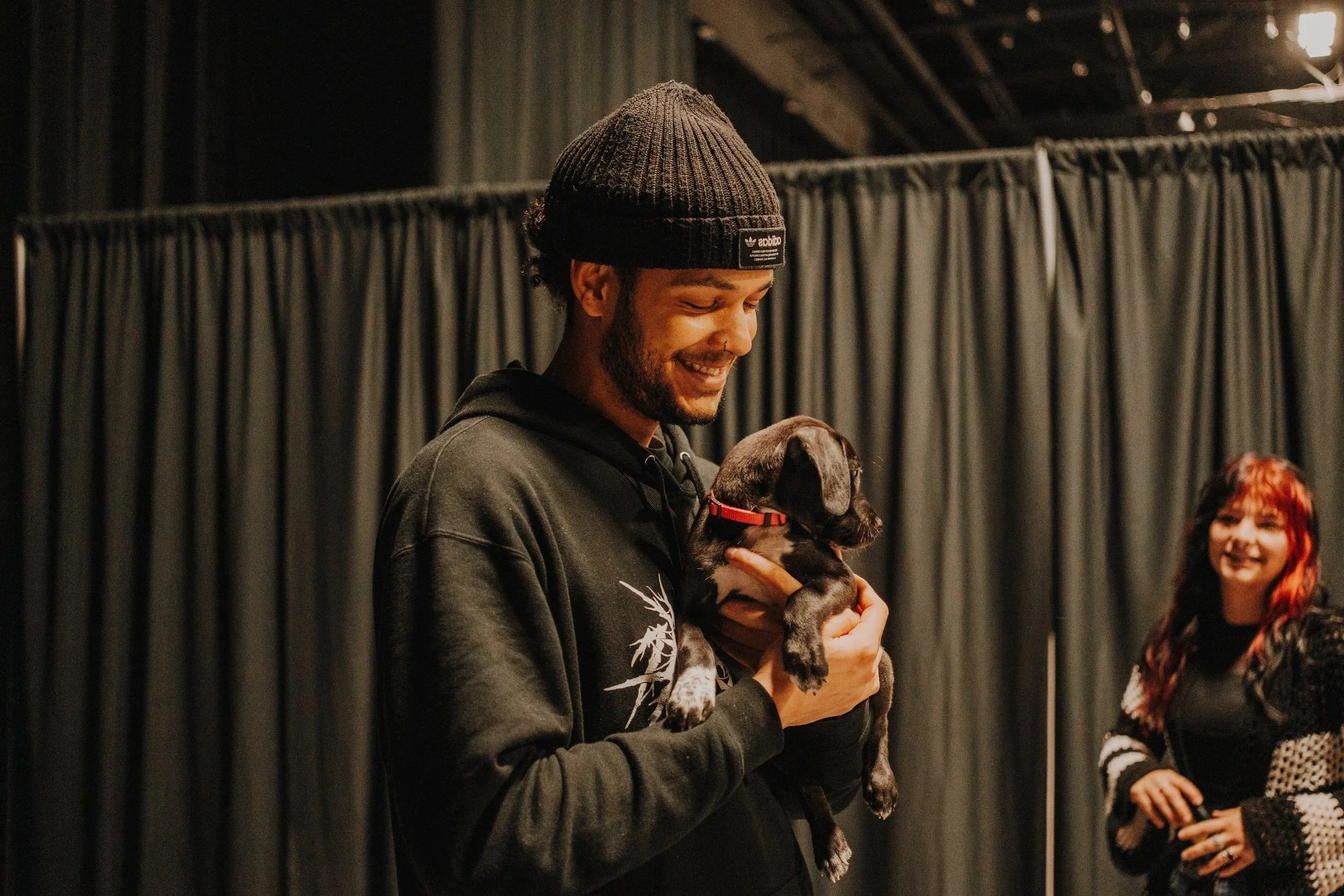 A man in a black beanie and hoodie holding a small black and gray puppy, smiling in an indoor setting with dark curtains in the background. A woman with red hair and a black and white sweater is smiling in the background.