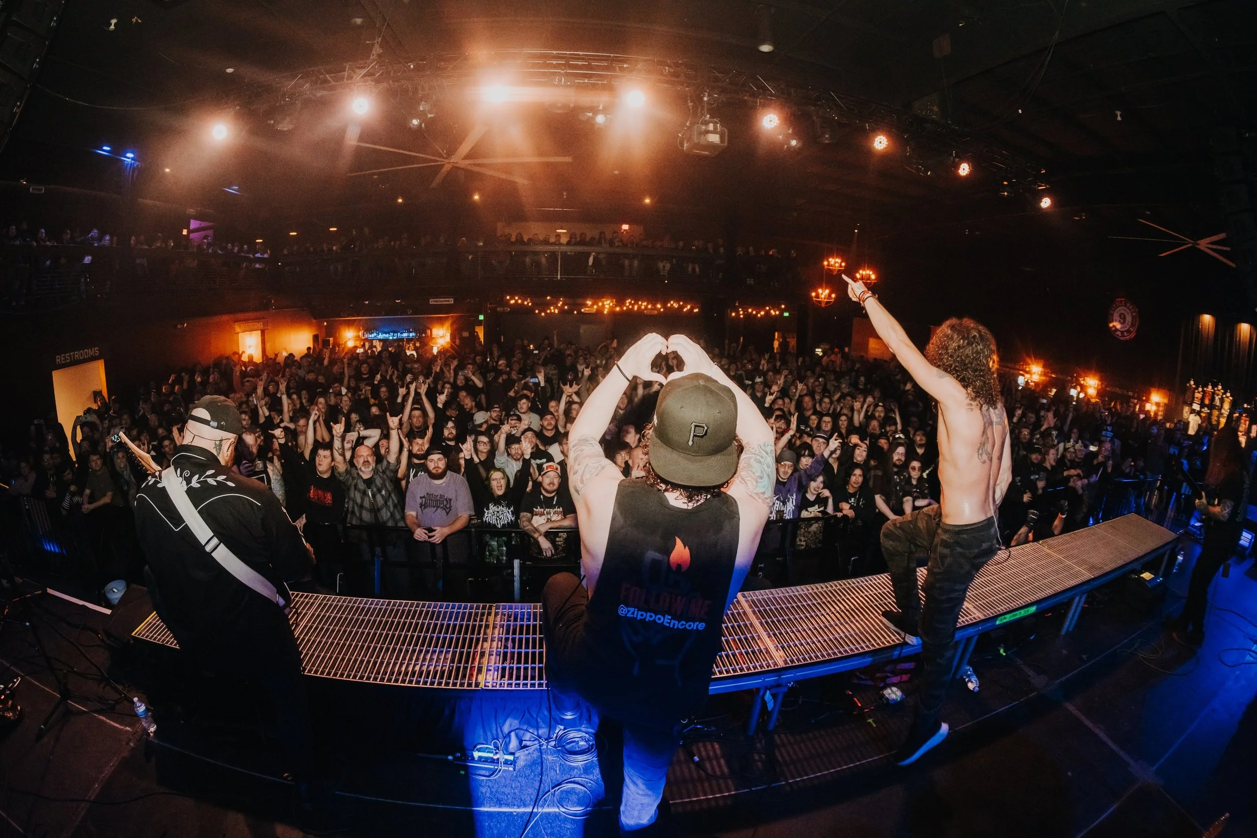 Musicians on stage performing in front of a crowd at a concert venue, with the audience raising their hands and forming heart shapes.