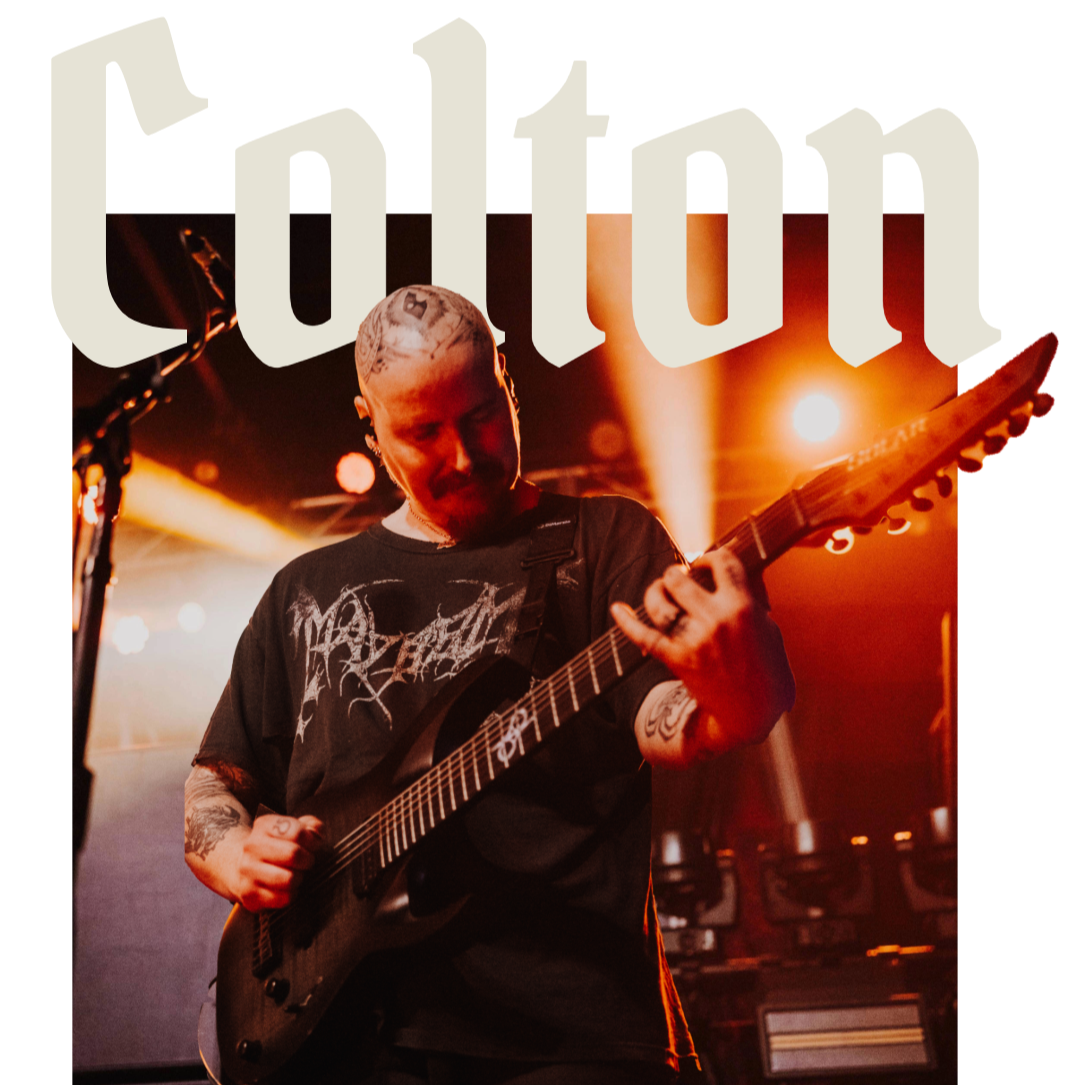 A musician with tattoos, wearing a black t-shirt, playing an electric guitar on stage with orange and yellow stage lighting.