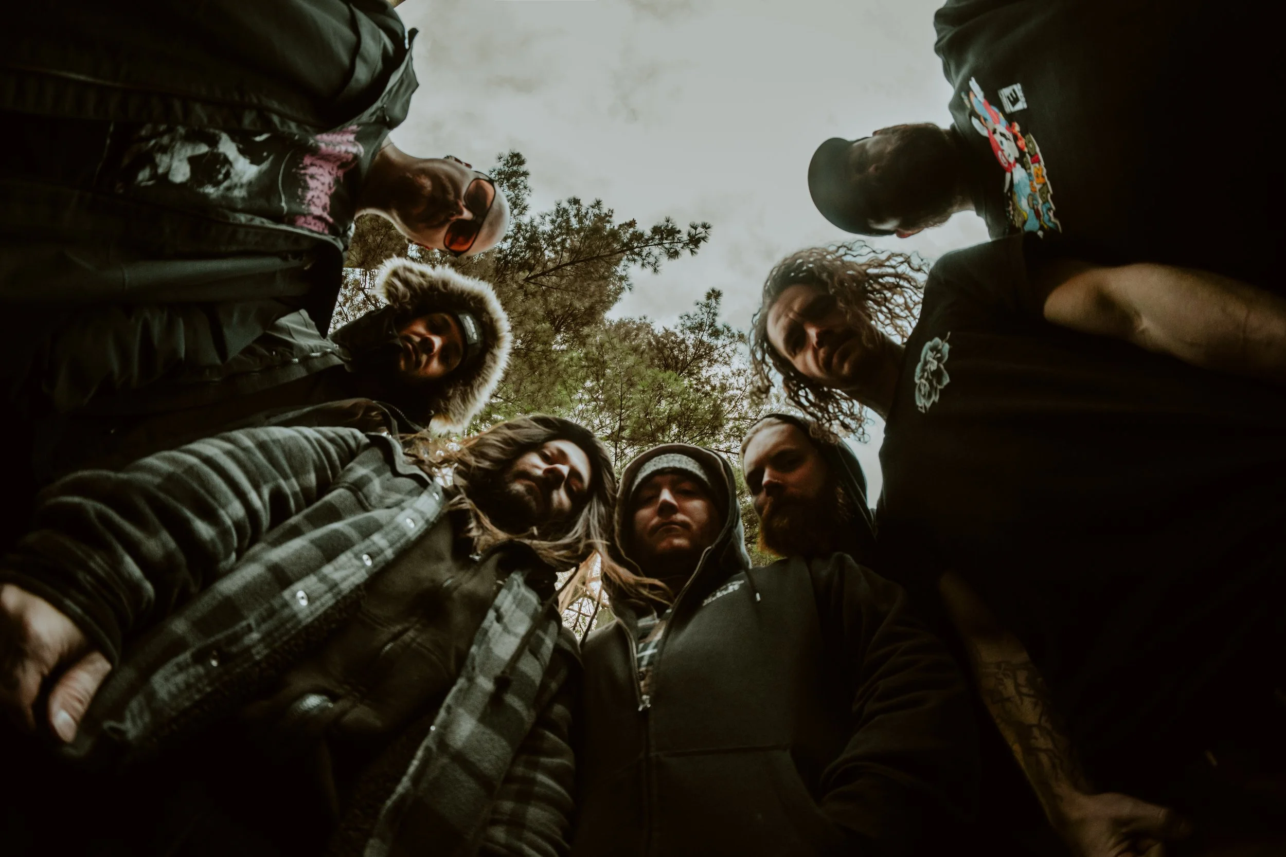 Group of seven people standing in a circle outdoors, looking down into the camera, with trees and cloudy sky overhead.