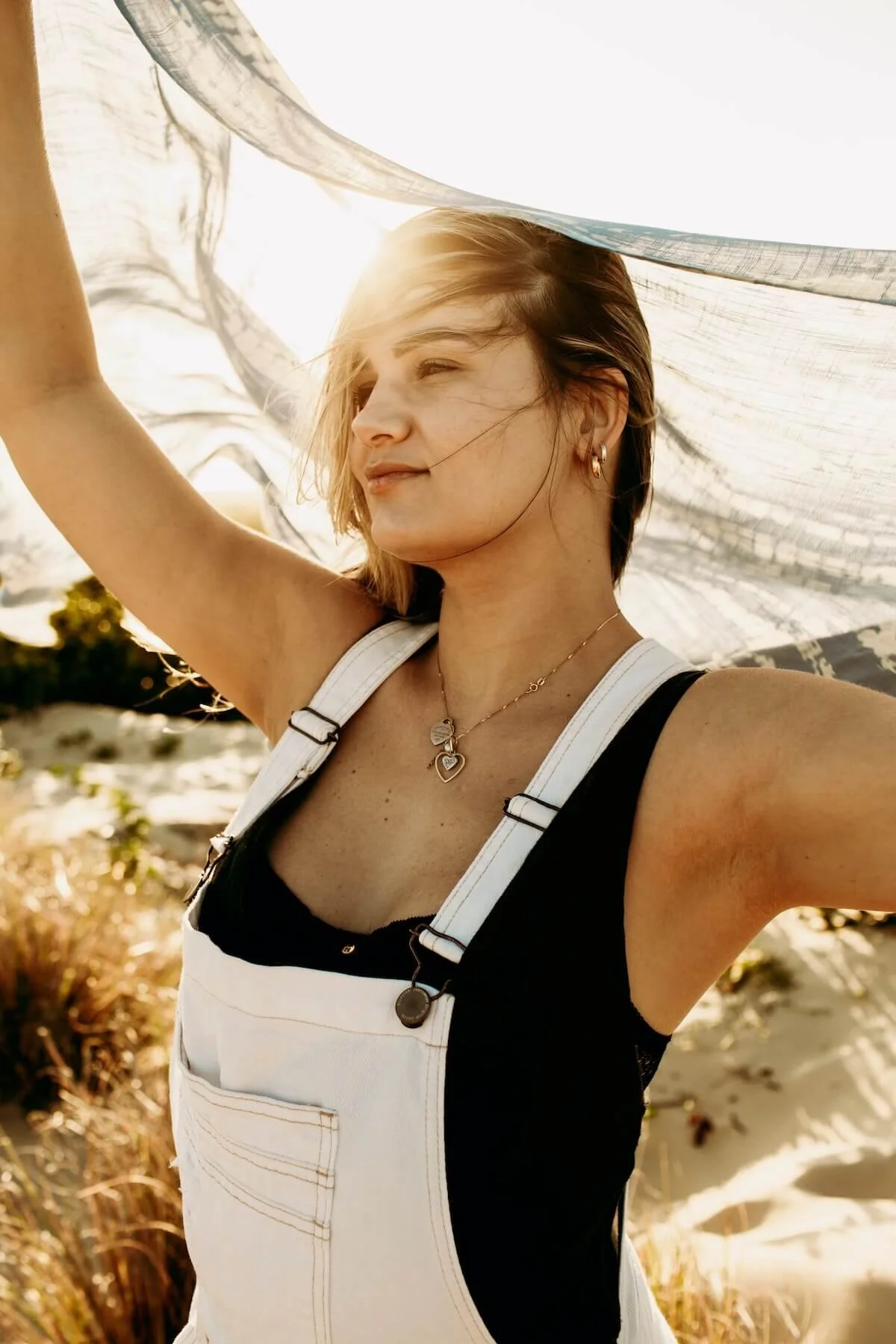 A woman with blonde hair is smiling with her eyes closed, holding a fabric above her head, at a beach during sunset, wearing white overalls with a black top underneath, gold jewelry, and earrings.