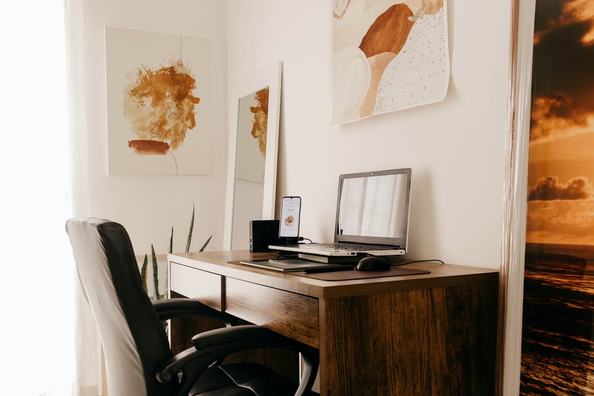 An organized home office with a wooden desk holding a laptop, mouse, smartphone, and a small black speaker. Three black office chairs are positioned around the desk. The background features white walls adorned with abstract art and a landscape painting of a sunset over clouds, a mirror, and a green plant with long leaves.