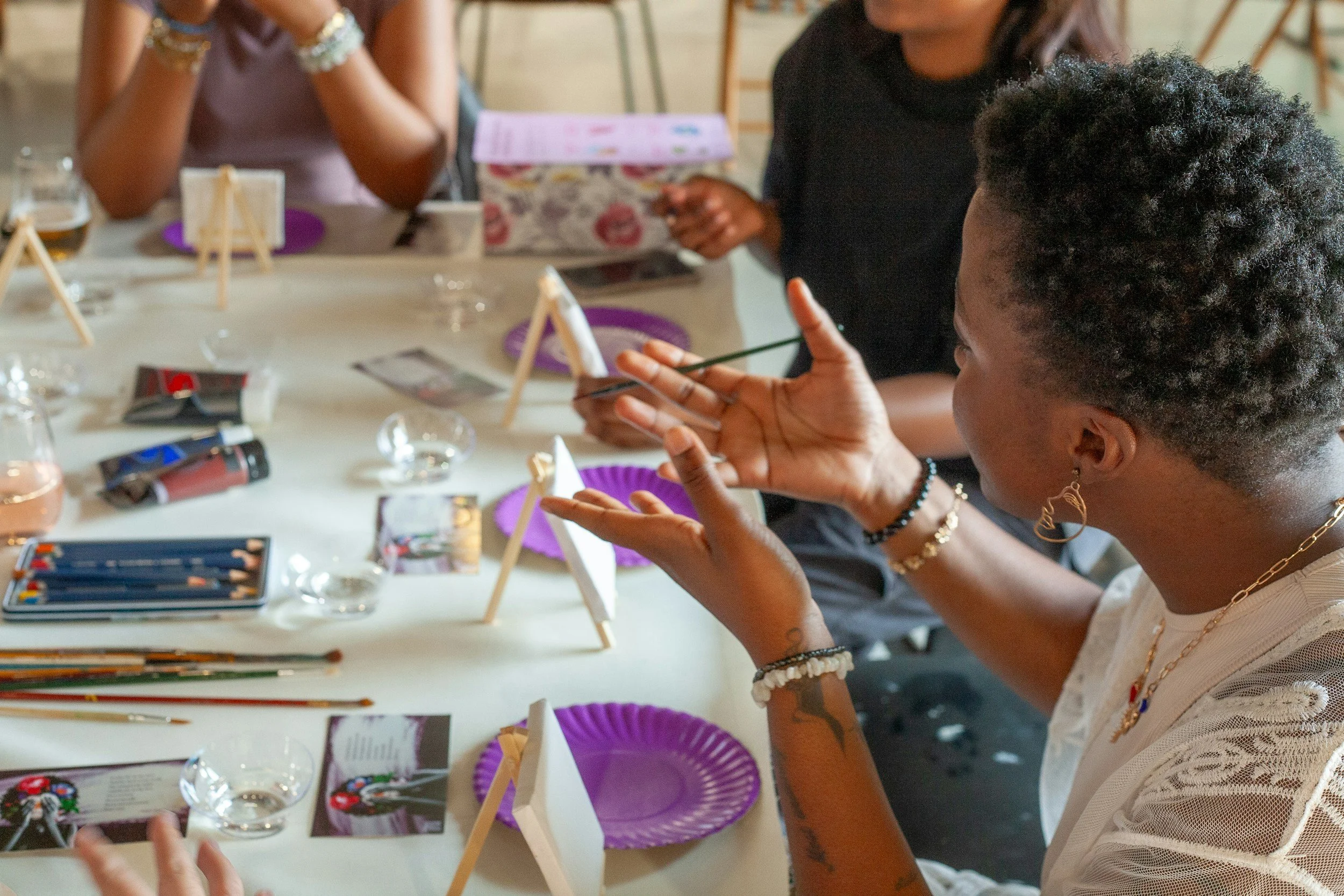 Three women sitting around a table with art supplies, including colored pencils, small easels, and decorative items, engaging in a creative activity or craft session.