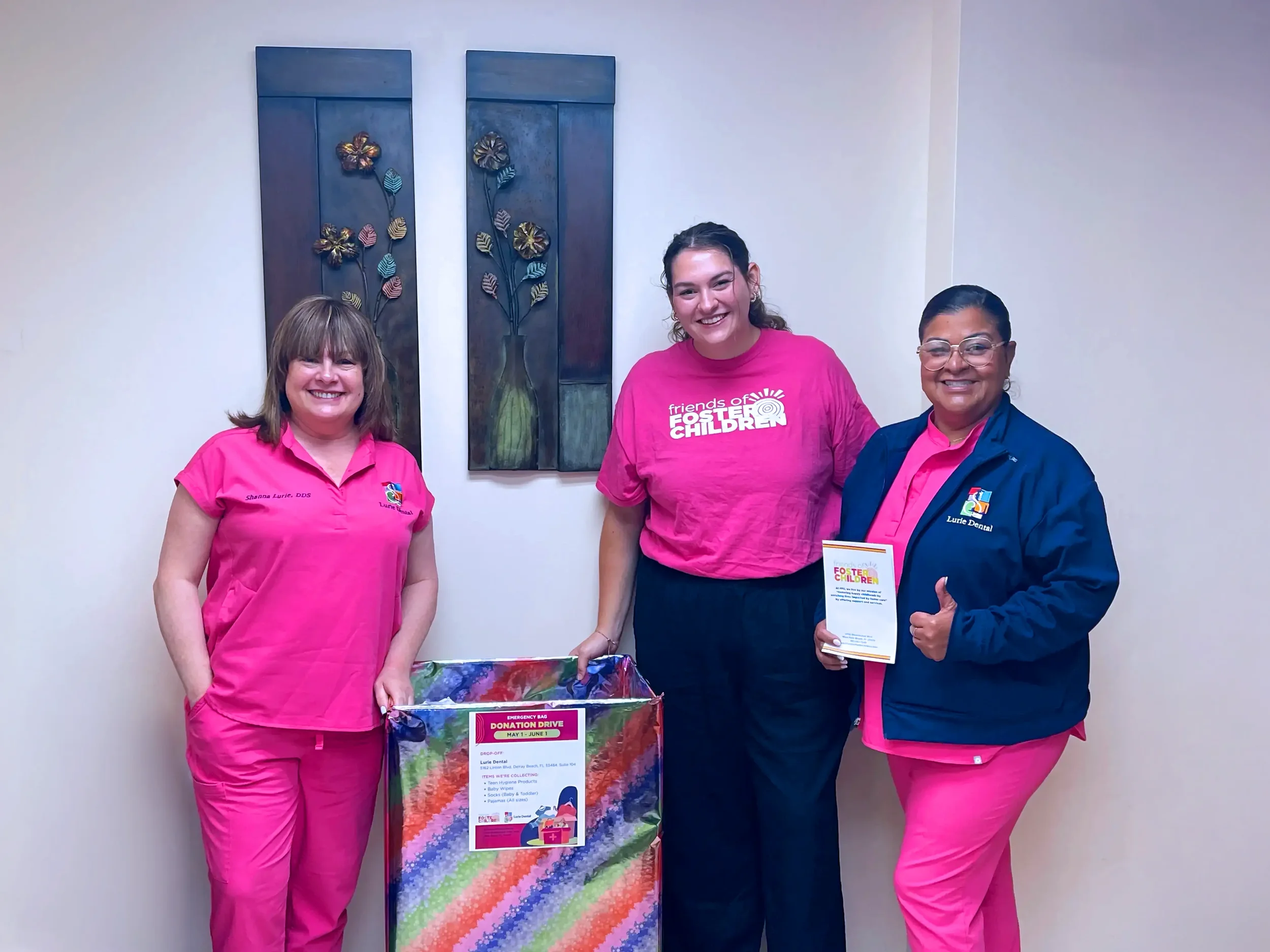 Three women standing in front of a wall, each wearing a pink or navy jacket and pink scrubs, smiling, holding a colorful donation bag and a flyer, in an indoor setting with decorative art on the wall behind them.