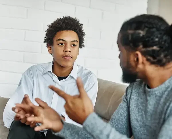 A young man with curly hair and a white shirt listens as an adult man with braids and a gray sweater speaks and gestures during a serious conversation indoors.