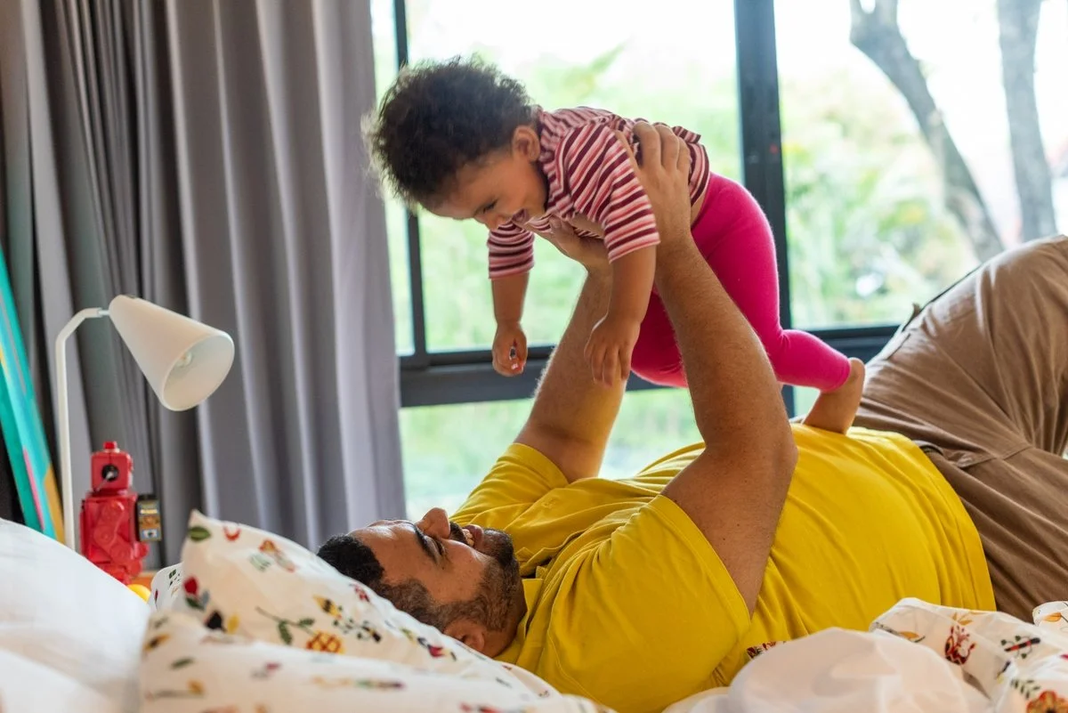 A man lying on a bed with a pillow, holding a young girl above him, both smiling with a large window behind them showing greenery outside.