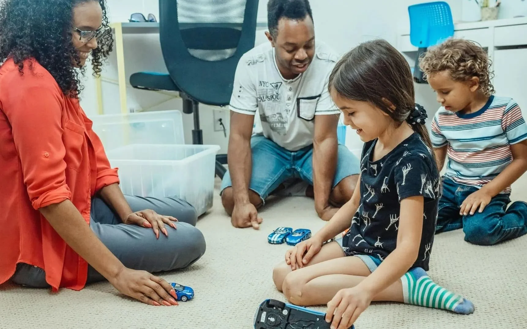A group of four people, two children and two adults, playing with toy cars on the floor of a room. The children are sitting on the carpet, and the adults are kneeling or sitting nearby, engaging with the children and the toys.