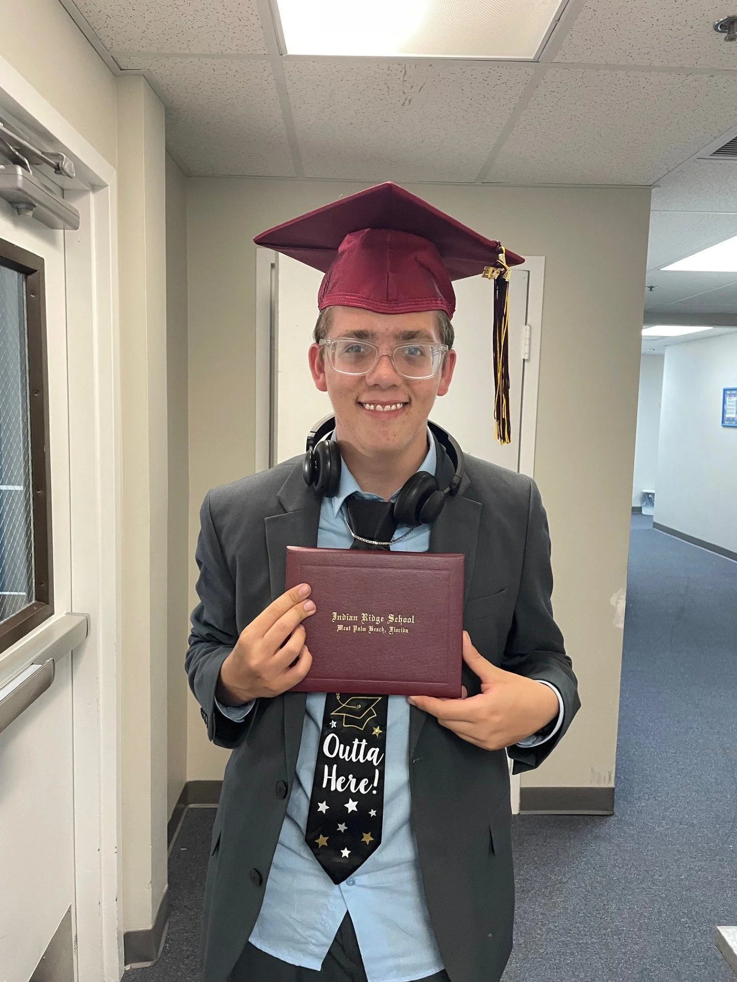 A young man in a suit with a maroon graduation cap and clear glasses holding a diploma cover that reads 'Indian Ridge School, West Palm Beach, Florida'. He is smiling and wearing headphones around his neck.