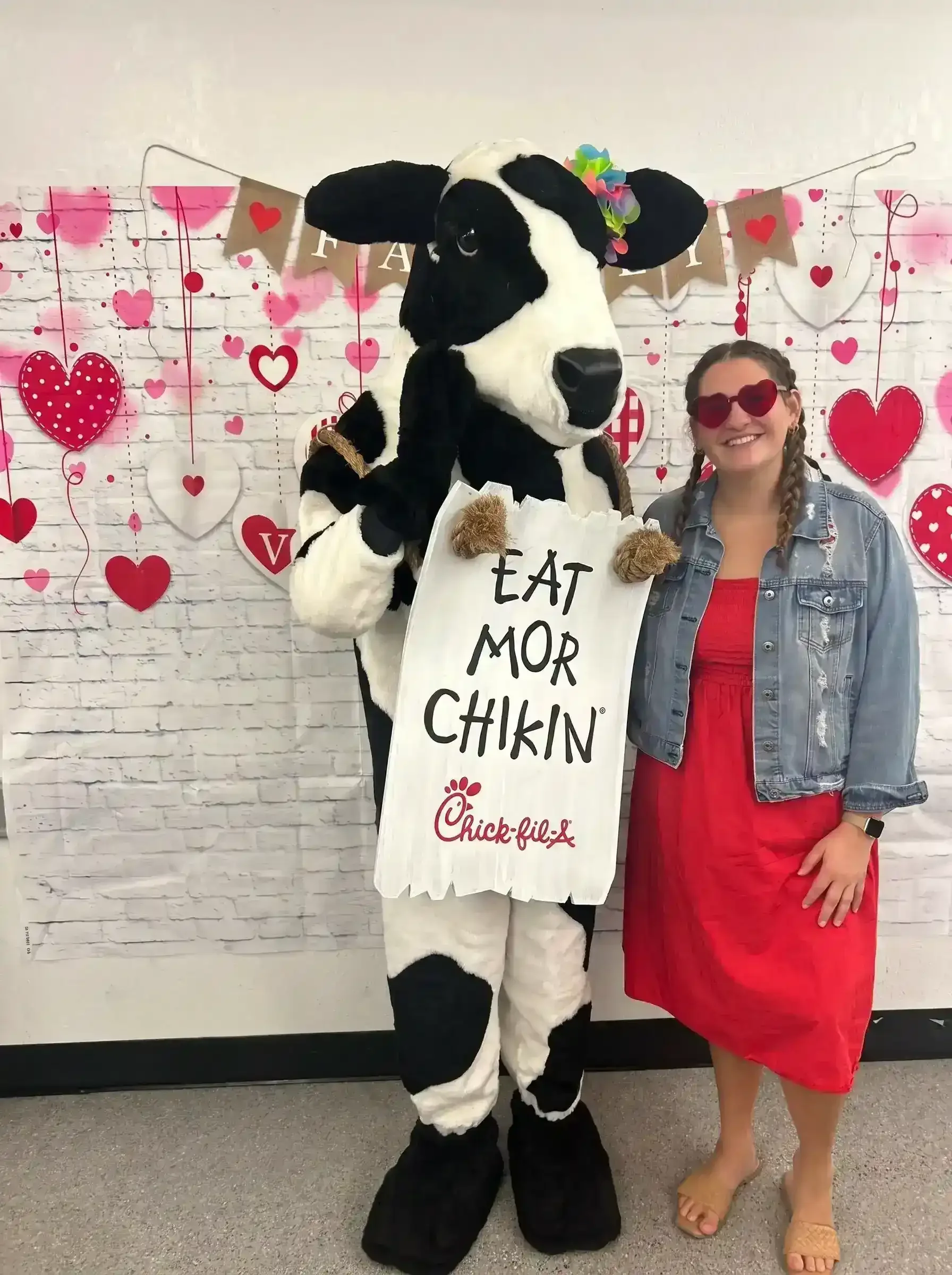 Woman standing with a person in a cow costume in front of a Valentine-themed backdrop, with hearts and banners. The woman on the right is wearing a denim jacket and a red dress