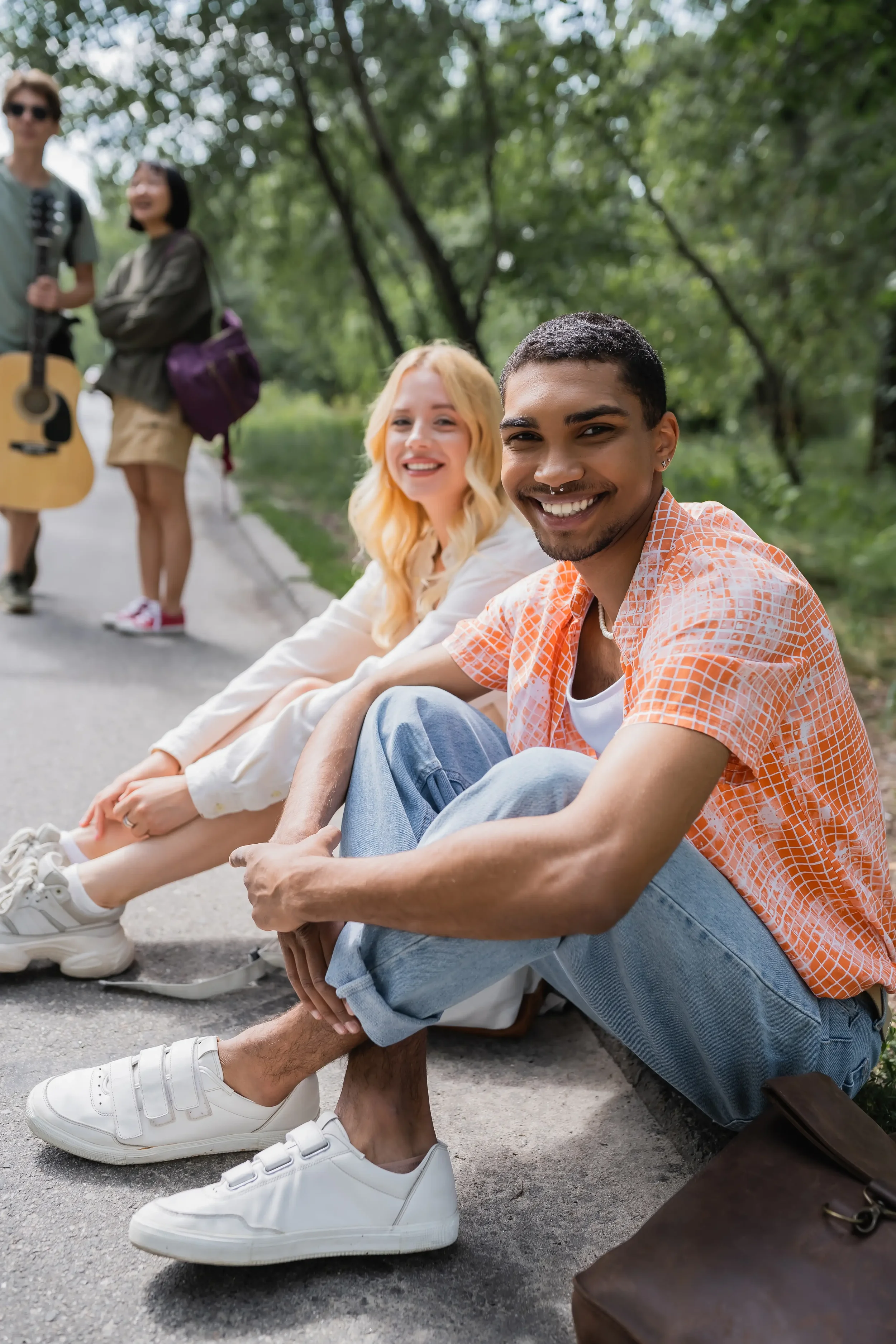 Two young adults sitting on a curb among trees, smiling at the camera, with two more people standing in the background, one holding a guitar.