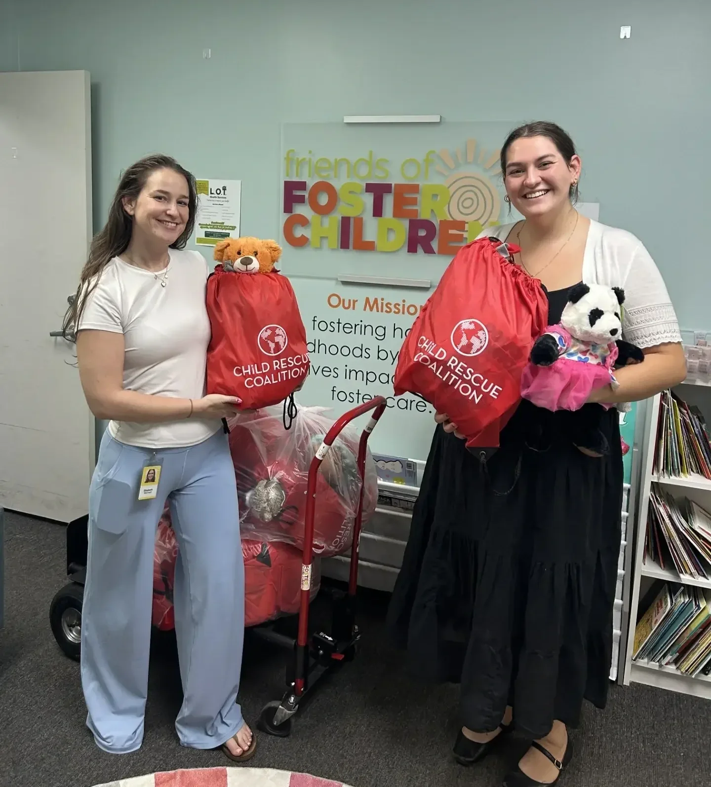 Two women smiling and holding red gift bags with stuffed animals, standing in front of the Freinds of Foster Children logo