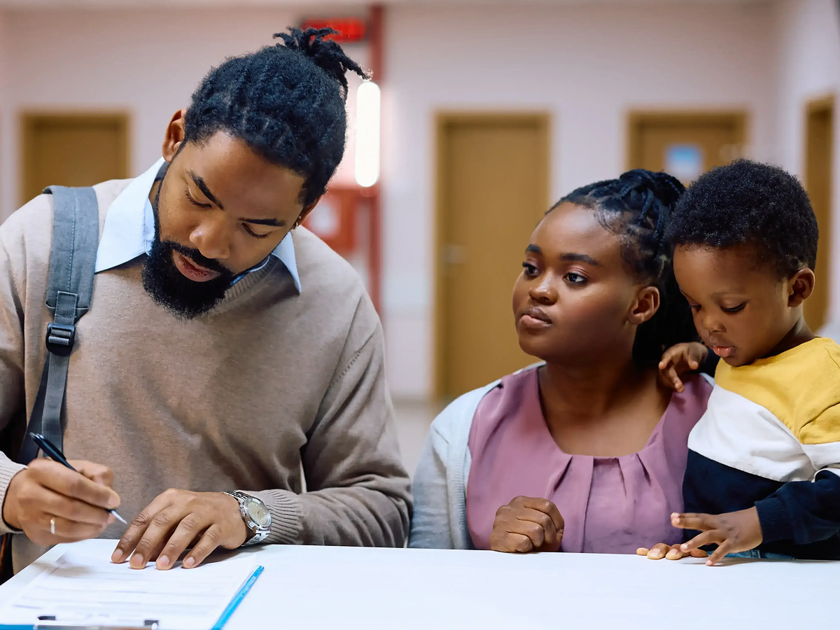 A man with a backpack signing a document at a table, with a woman and a young boy watching.