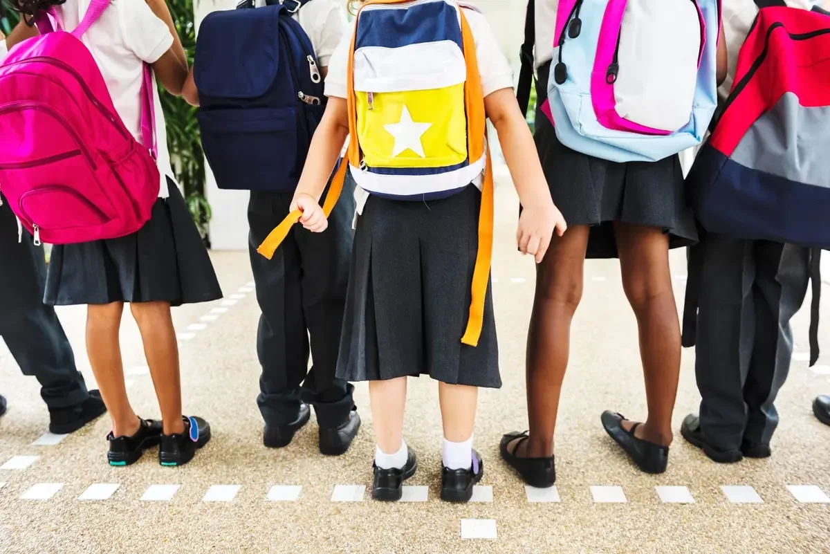 Children standing in a row with colorful backpacks, wearing school uniforms, in an indoor setting.