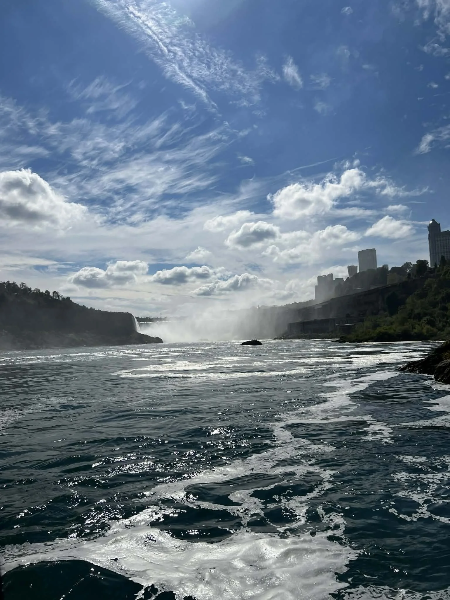 View of a river with waterfall in the distance, surrounded by hills, with a clear blue sky and scattered clouds overhead. Part of a city skyline is visible on the right side of the image.