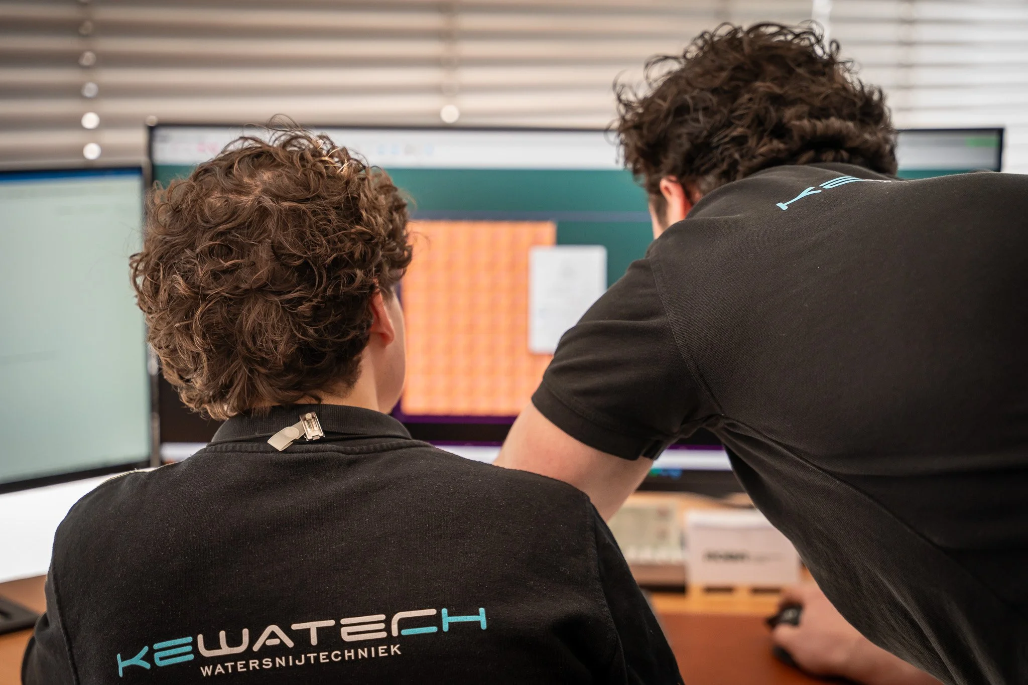 Two men with curly hair working together at a computer station with multiple monitors in an office setting, wearing black shirts with a logo that reads 'KEWATECH WATERSNIJTECHNIEK'.