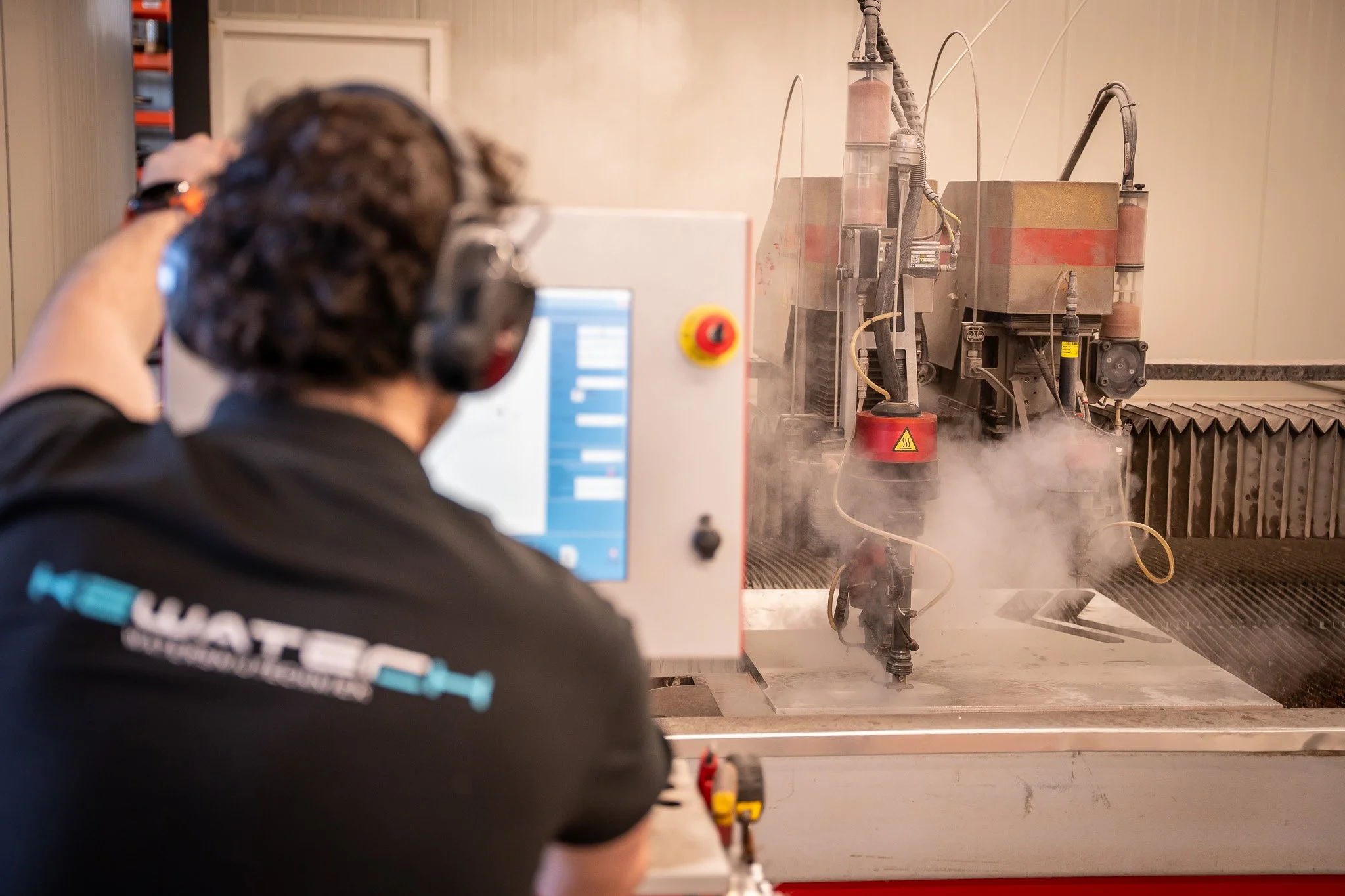 A person wearing safety ear protection and a shirt with a logo watches a CNC machine in operation, with smoke or steam rising from the work area.