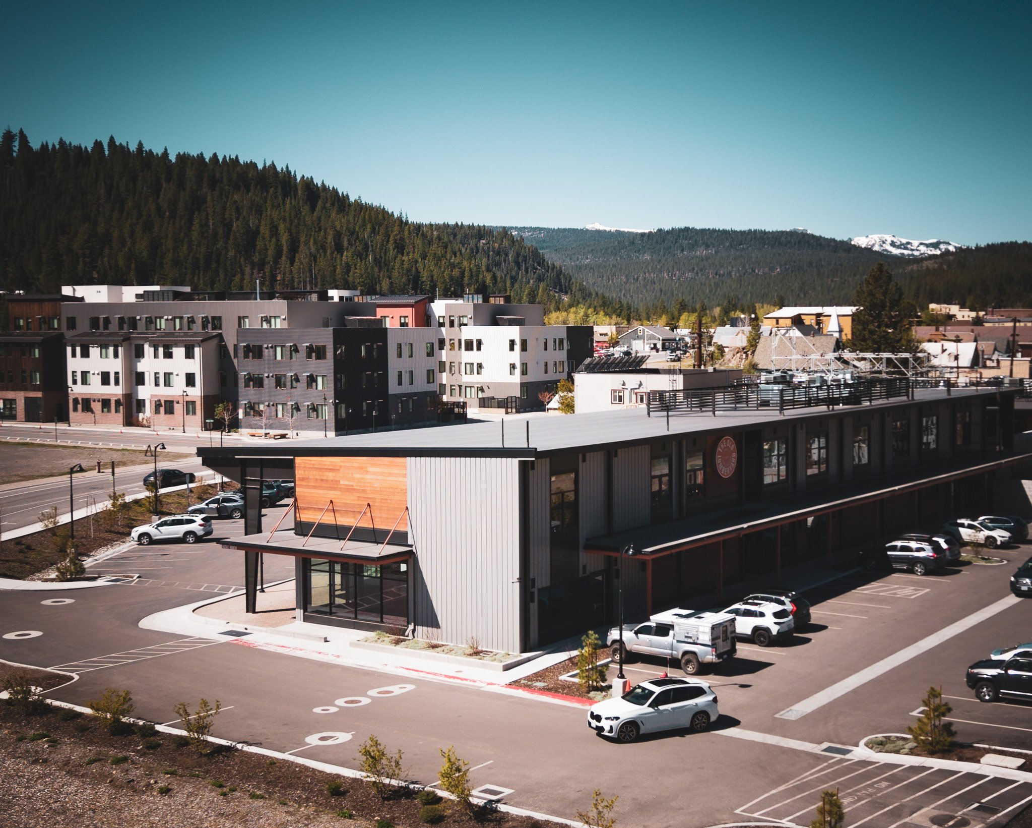 Cityscape of Truckee CA, featuring The Lumber Yard business plaza with modern residential and commercial buildings, parking lot with cars, lush green forested mountains in the background, and a clear blue sky.
