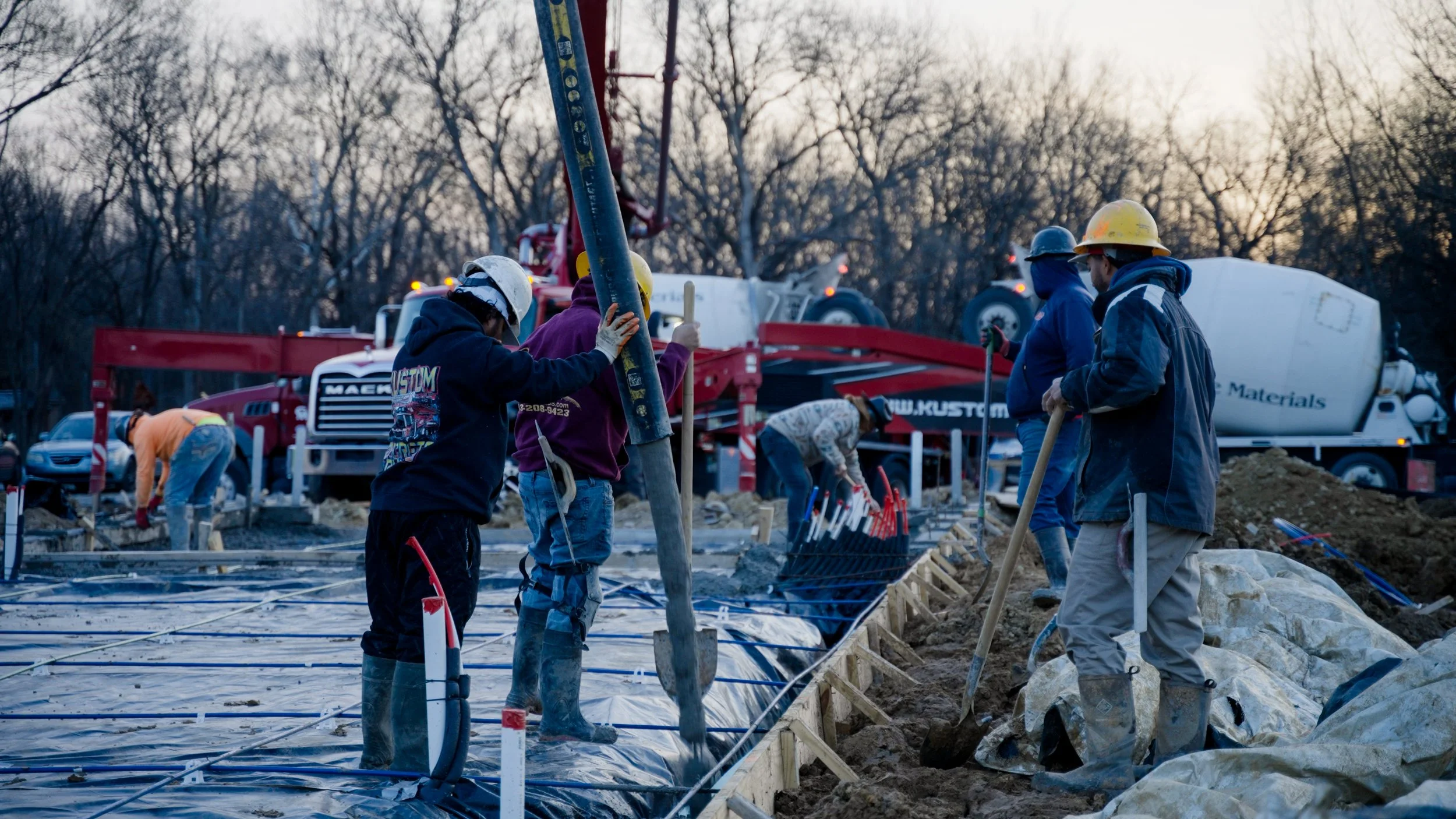 Construction workers in hard hats and boots working on a building foundation with concrete and rebar, with trucks and equipment in the background and trees around at dusk.