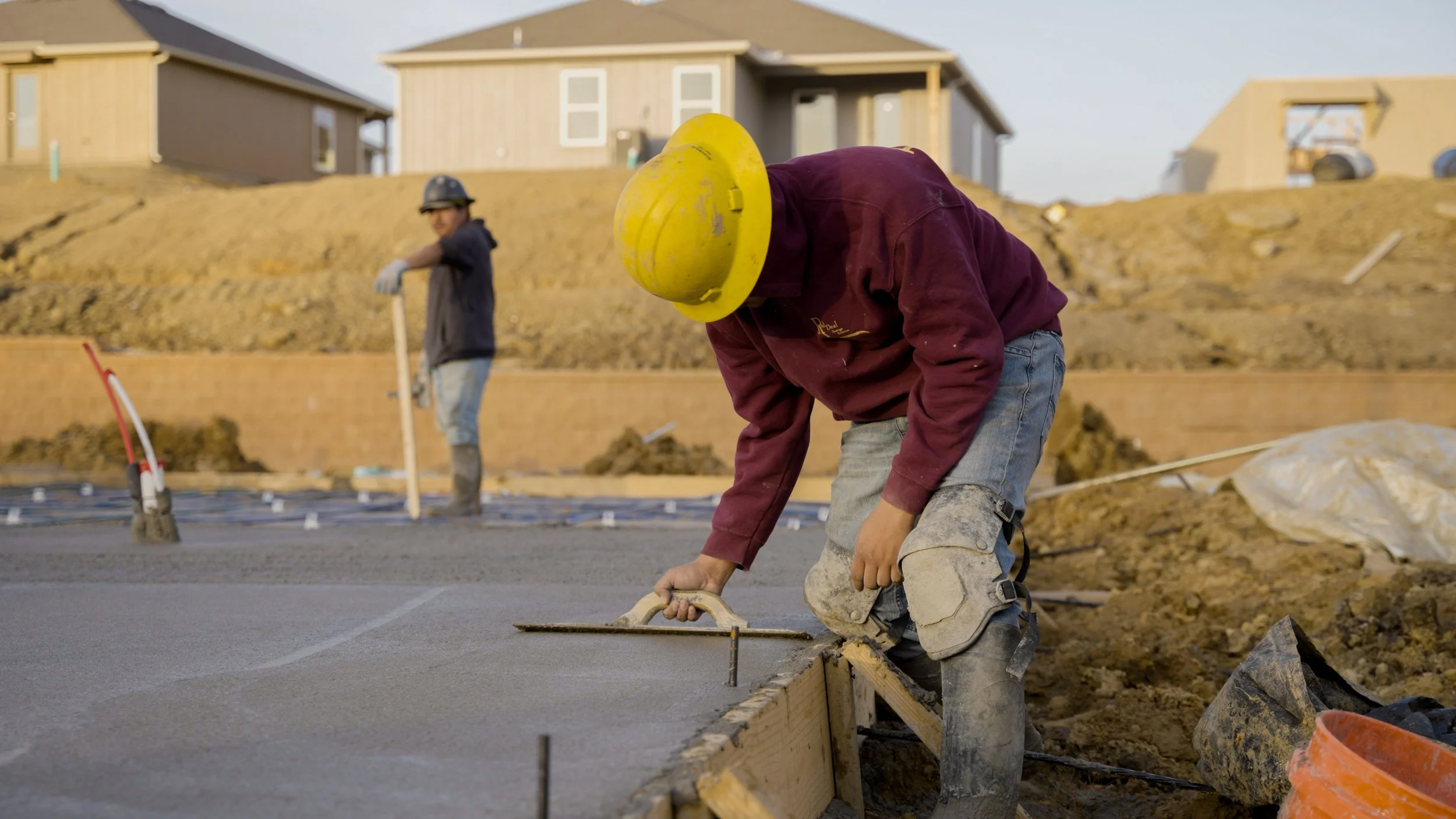 Construction workers pouring and leveling concrete on a building site, wearing safety helmets and gloves, with houses in the background.