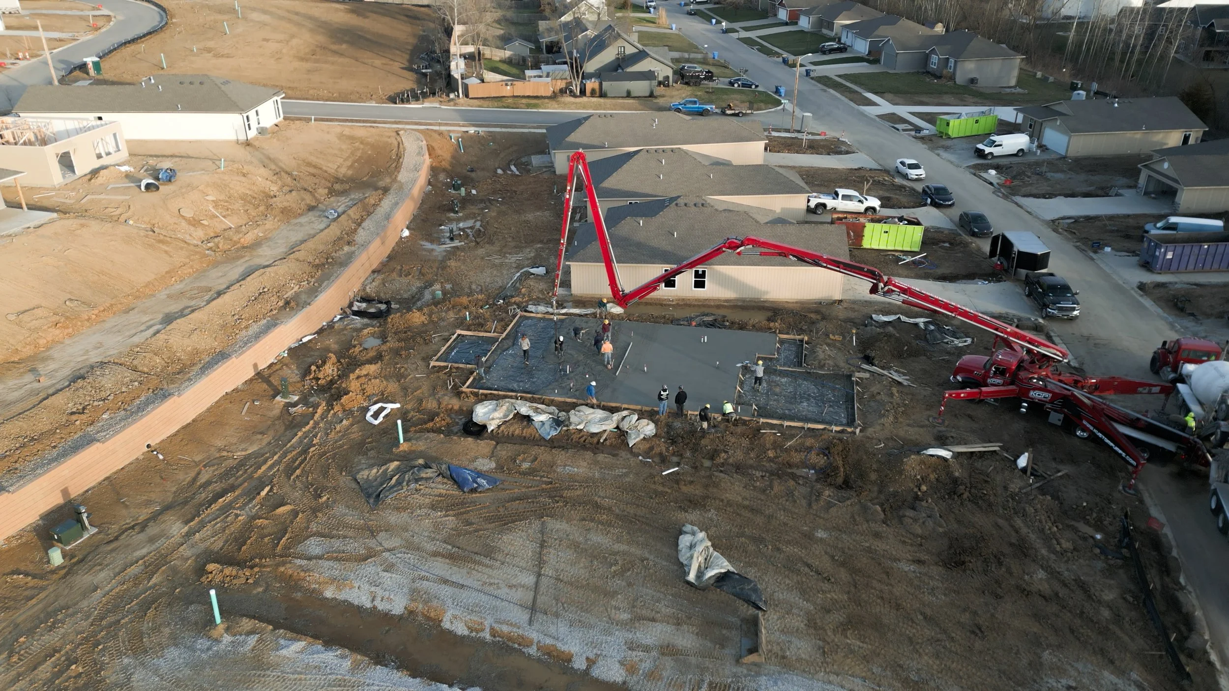 Construction site with workers pouring concrete for building foundation, a concrete pump truck positioned nearby, residential houses in the background.