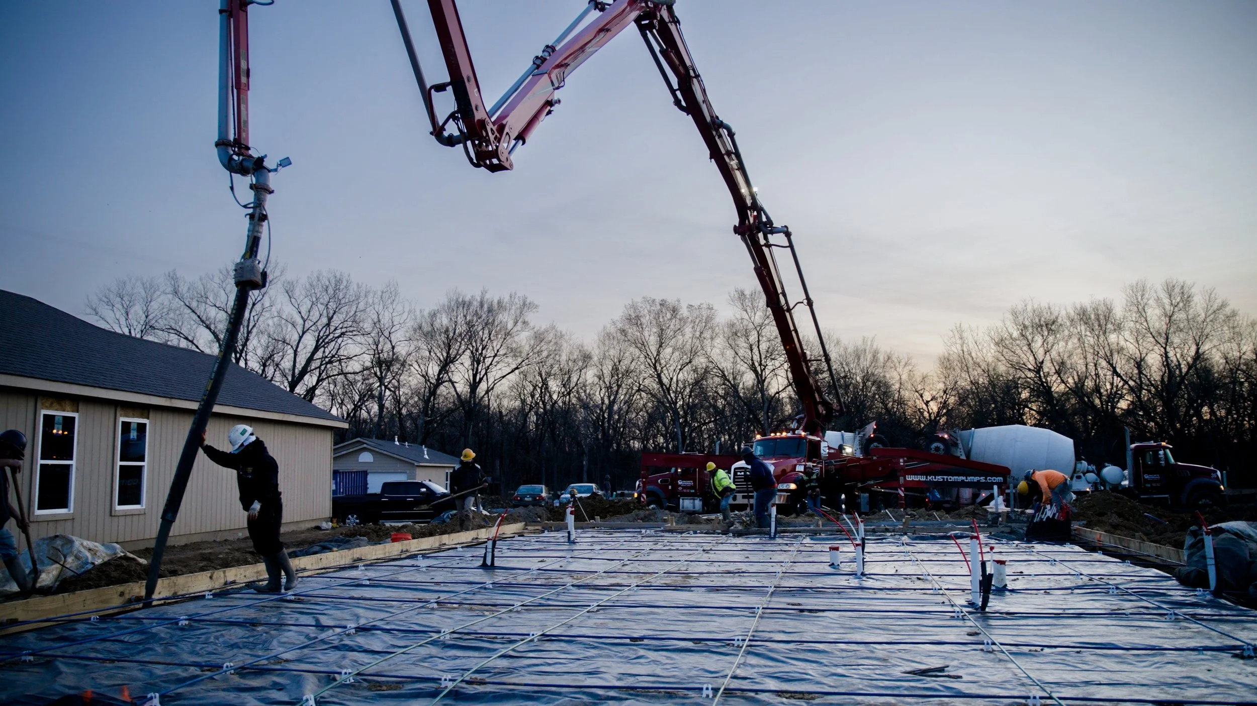 Construction workers pouring concrete on a building site during dusk, with a cement mixer truck and a concrete pump in the background.