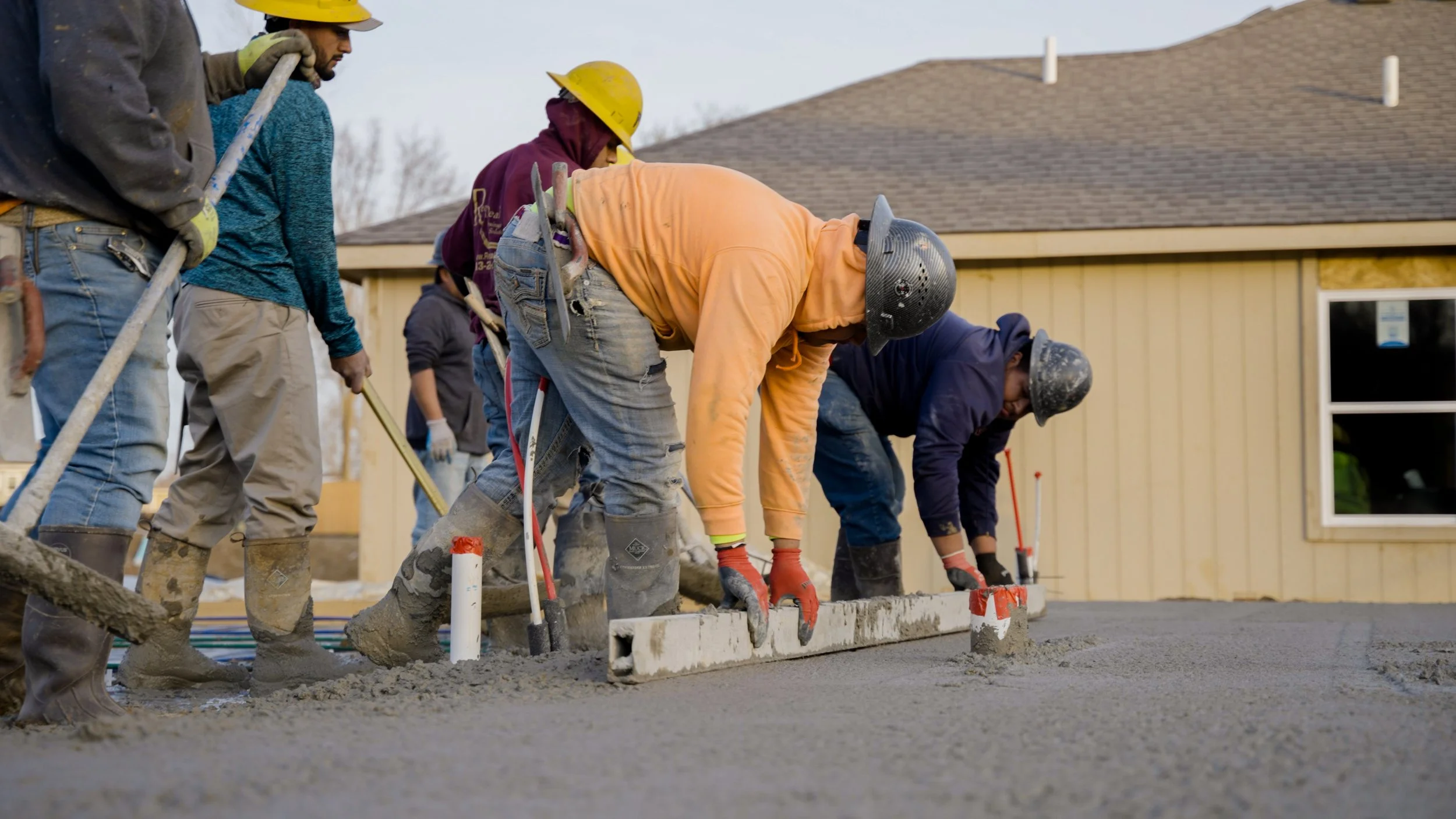 Construction workers pouring and leveling concrete on a new sidewalk or driveway, wearing safety helmets and gloves, in front of a beige house with a pitched roof.