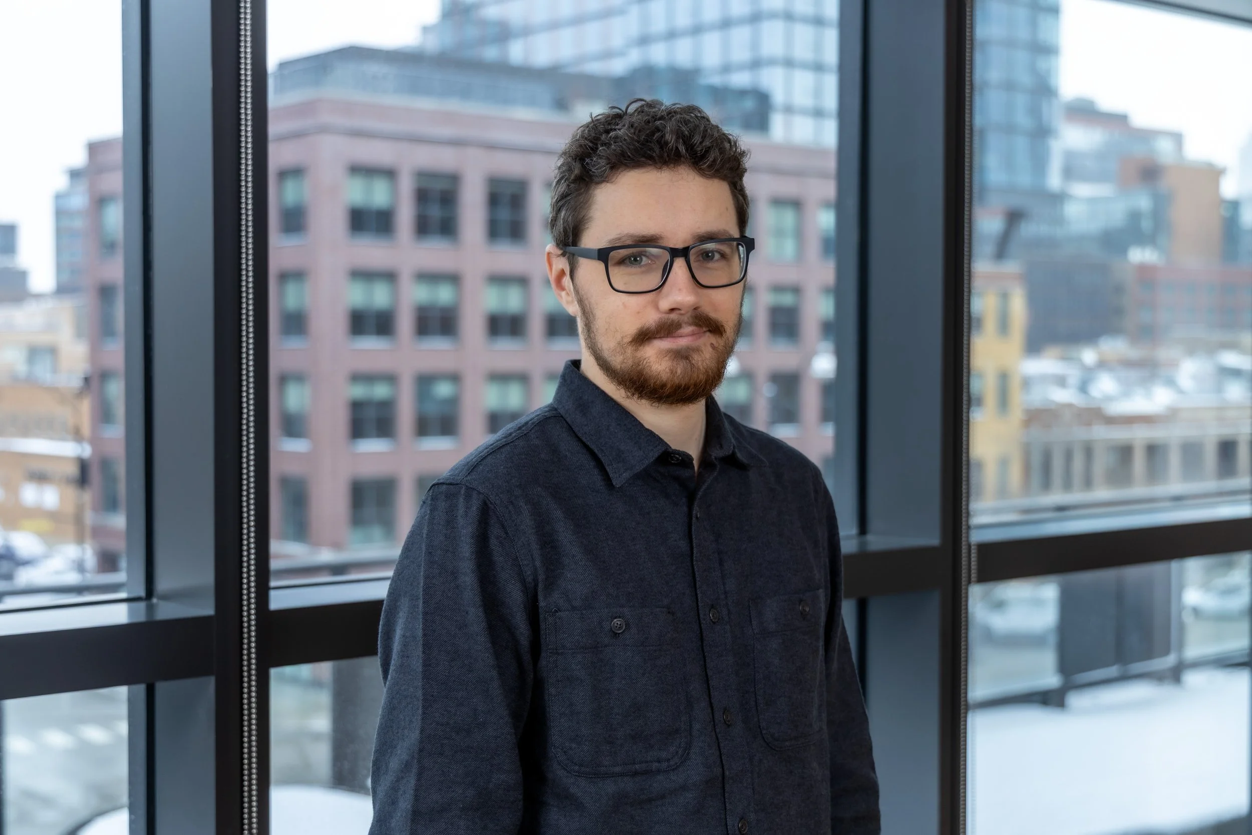 A man with glasses, a beard, and curly hair standing inside near large windows with an urban cityscape background.