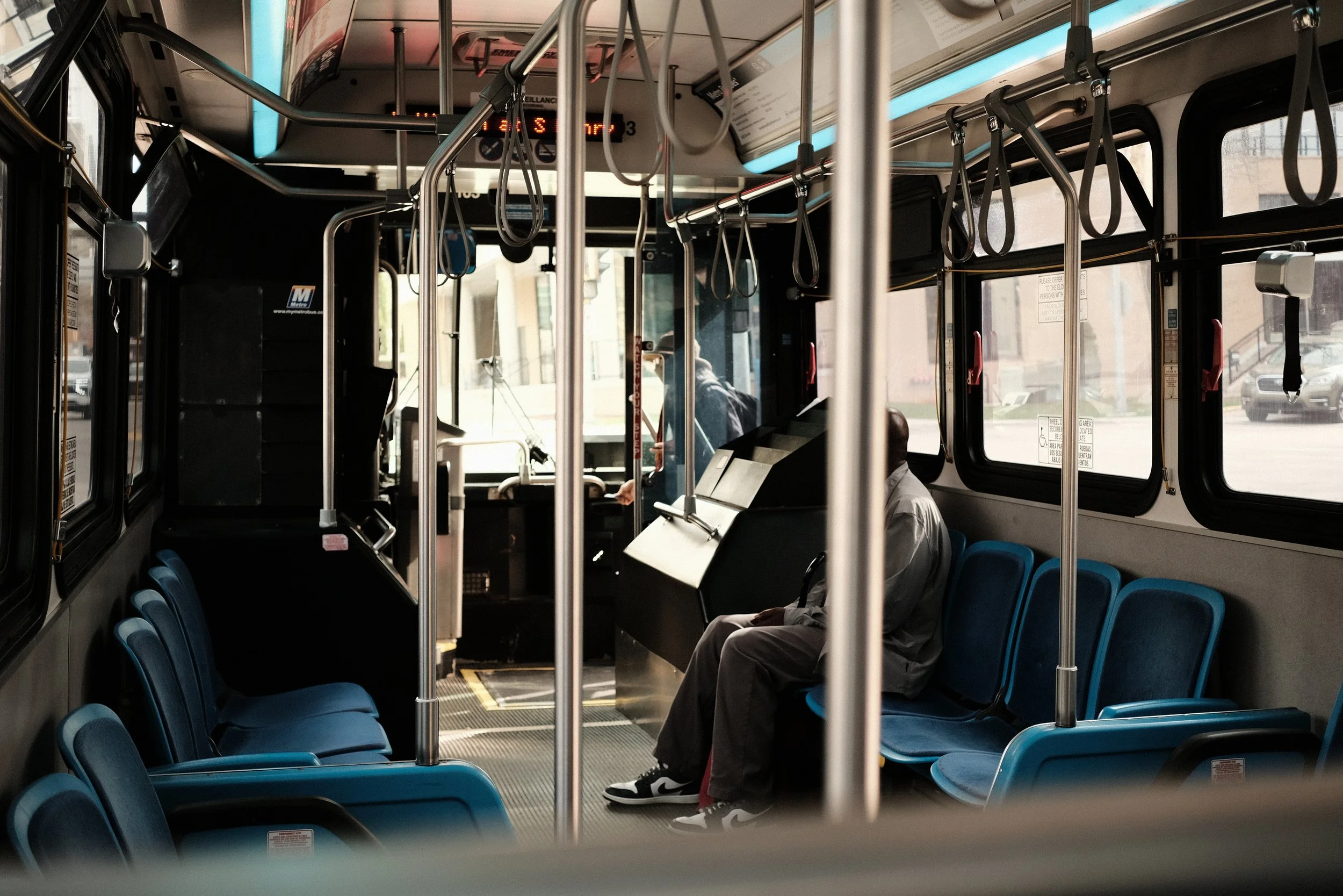 Inside a city bus with rows of blue seats, two passengers sitting, and the driver visible at the front. Handrails and straps hang from the ceiling, and windows reveal a city street outside.