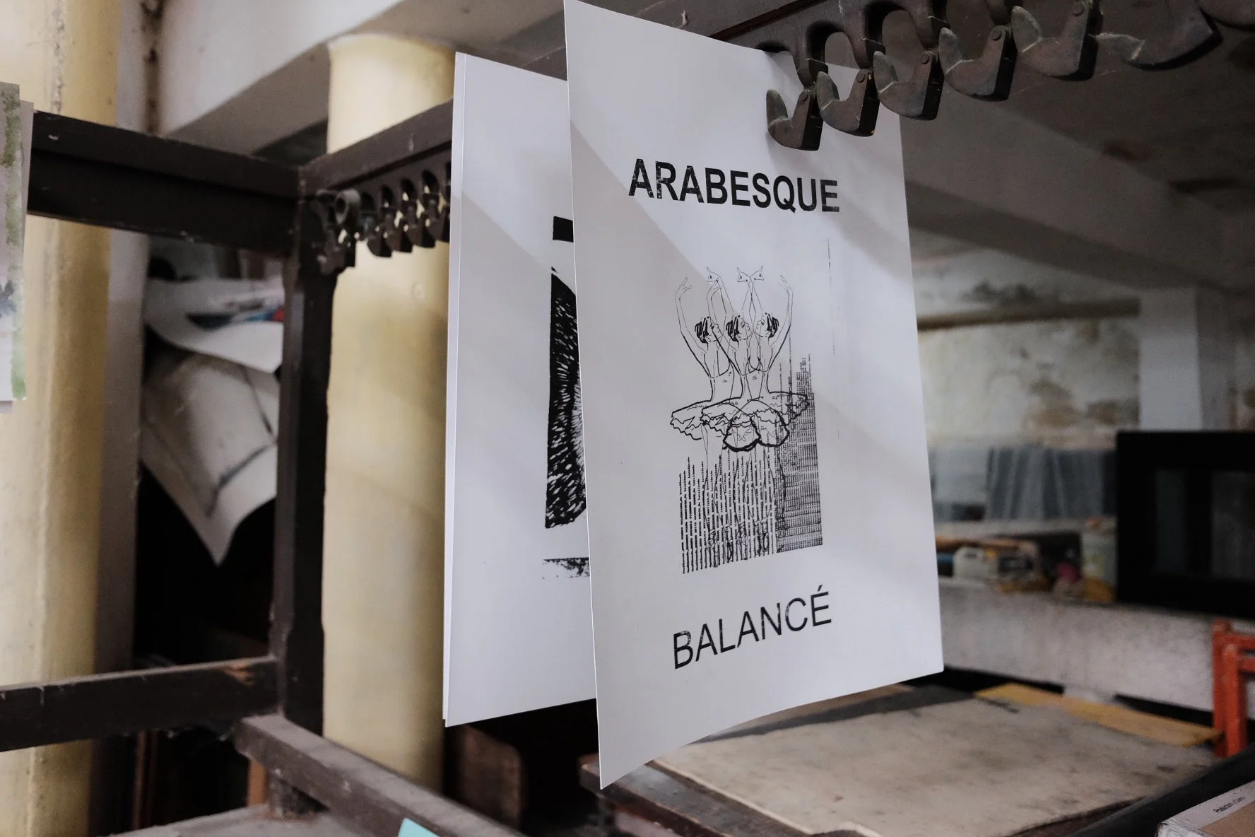 A printed page with the words 'ARABESQUE' and 'BALANCED' hanging on a metal rack, with a line drawing of ballet dancers in an arabesque pose.