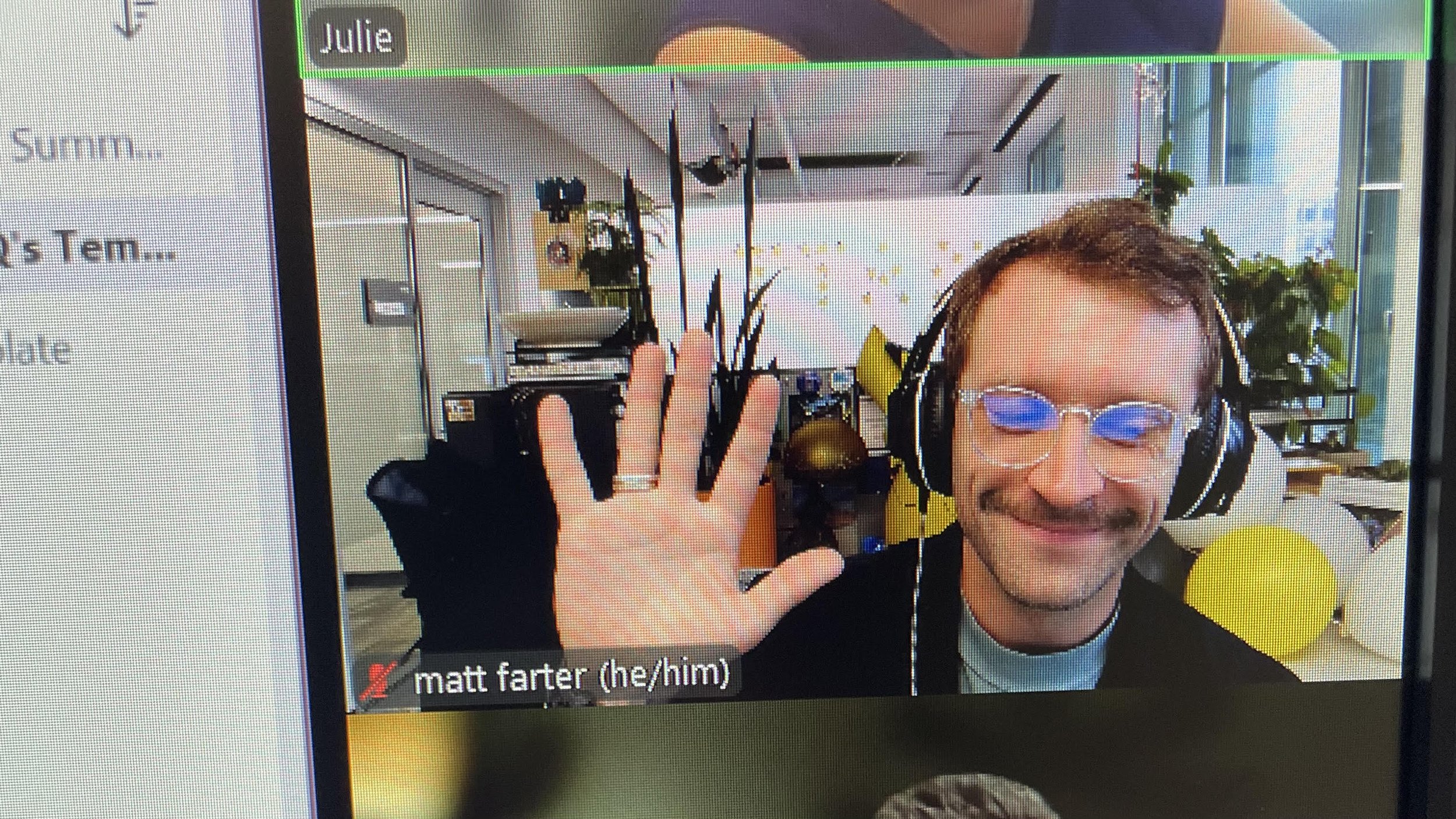 A man with glasses and a smile, wearing a headset, waving at the camera, in a brightly lit room with large windows, plants, and modern decor.