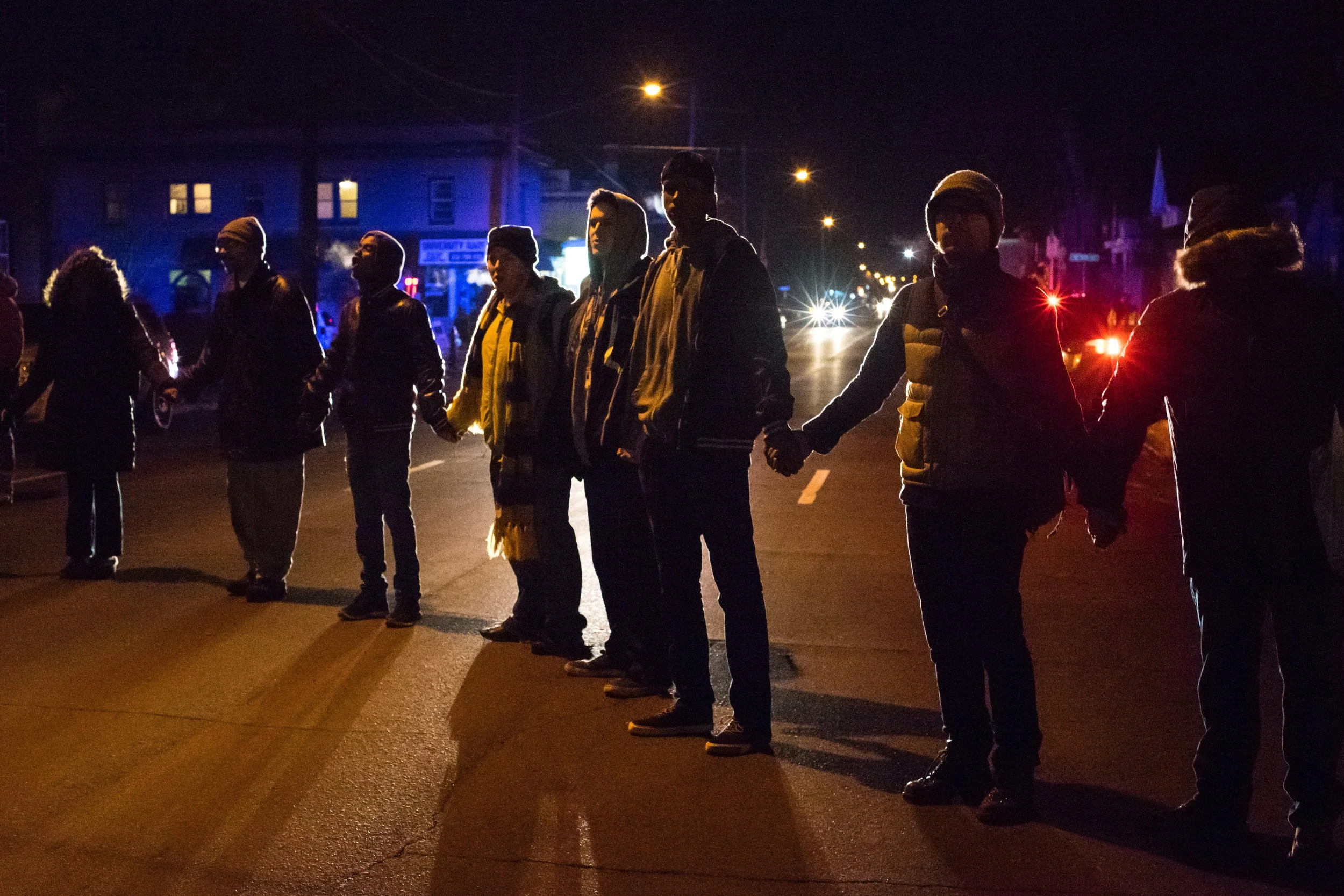 People holding hands in a line during nighttime on a street with police lights in the background.