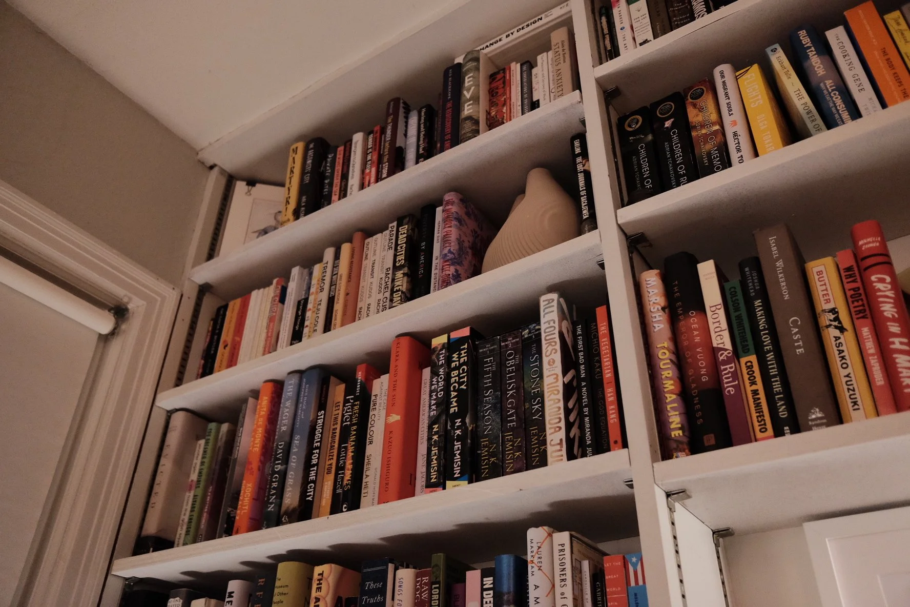 White bookshelves filled with a variety of books, some lying flat, with a framed picture and a ceramic vase on the middle shelf. The shelves are next to a window with a white frame.