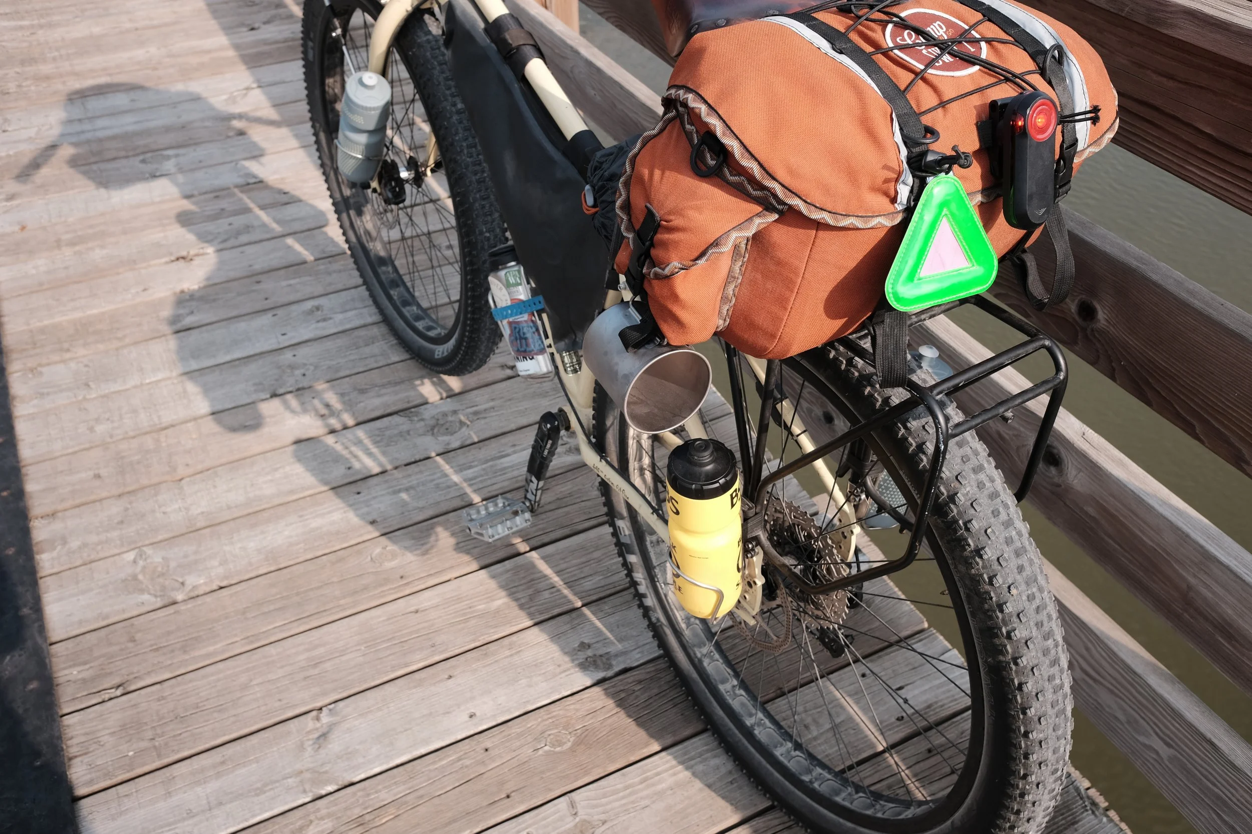 A bicycle on a wooden pier with an orange saddlebag attached to the rear rack. The bike has a water bottle in a holder, a rear light, a triangular reflector, and a black rack. The background shows water and part of the wooden railing of the pier.