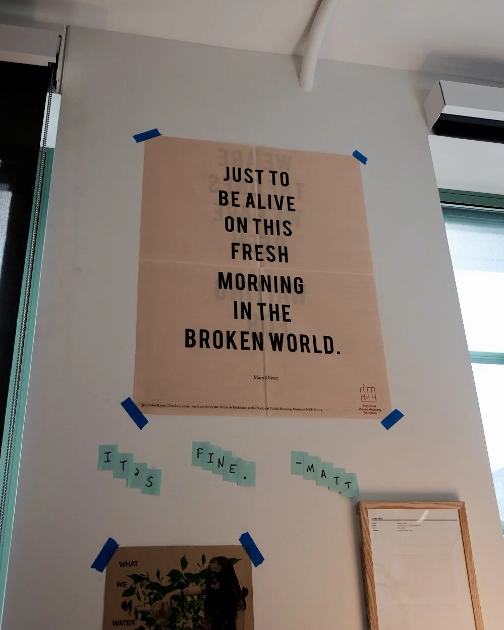 A wall with a large beige poster that says, "JUST TO BE ALIVE ON THIS FRESH MORNING IN THE BROKEN WORLD" by Mary Oliver. There are blue tape corners on the poster. Below are small, light blue paper notes spelling out "IT'S FINE. - MATT" and a partially visible framed document or picture.