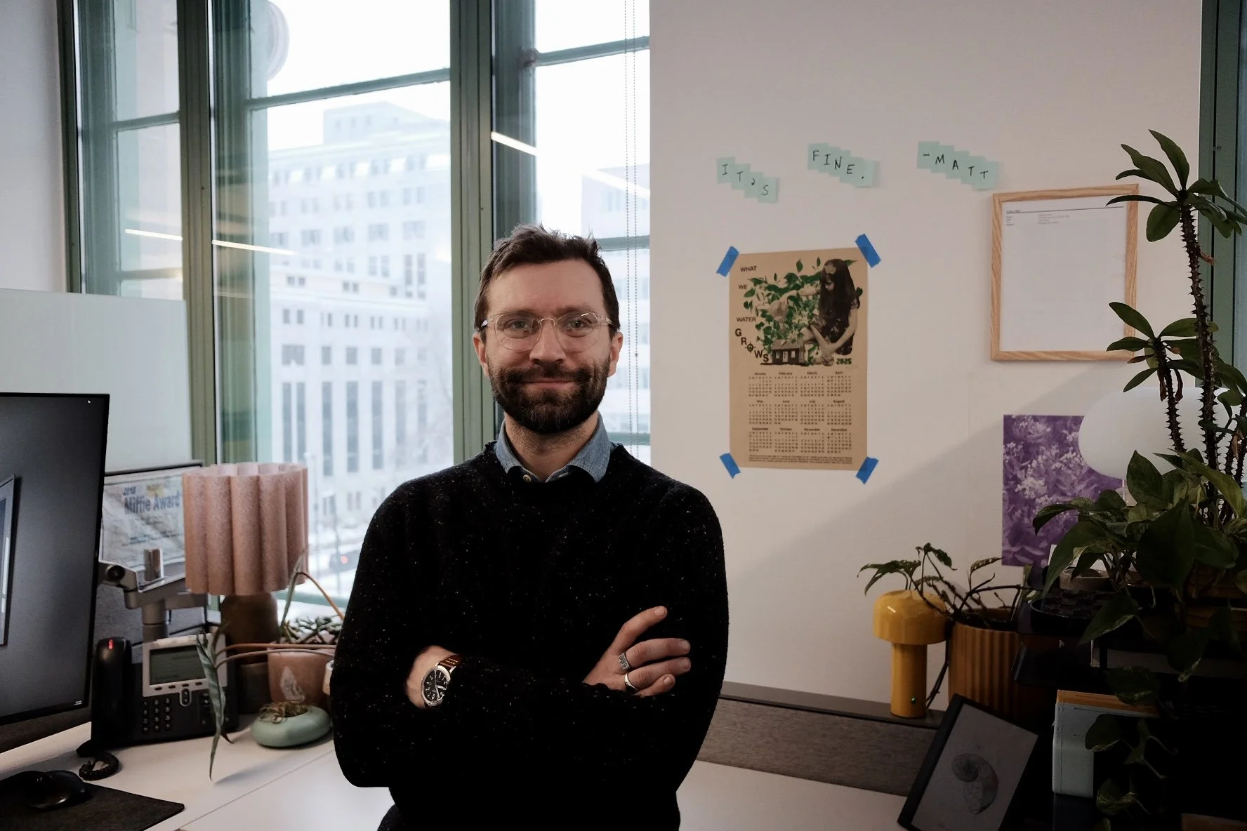 A man with glasses and a beard standing with arms crossed in an office, with large windows showing city buildings, a desk with a computer, and various plants and wall decorations.