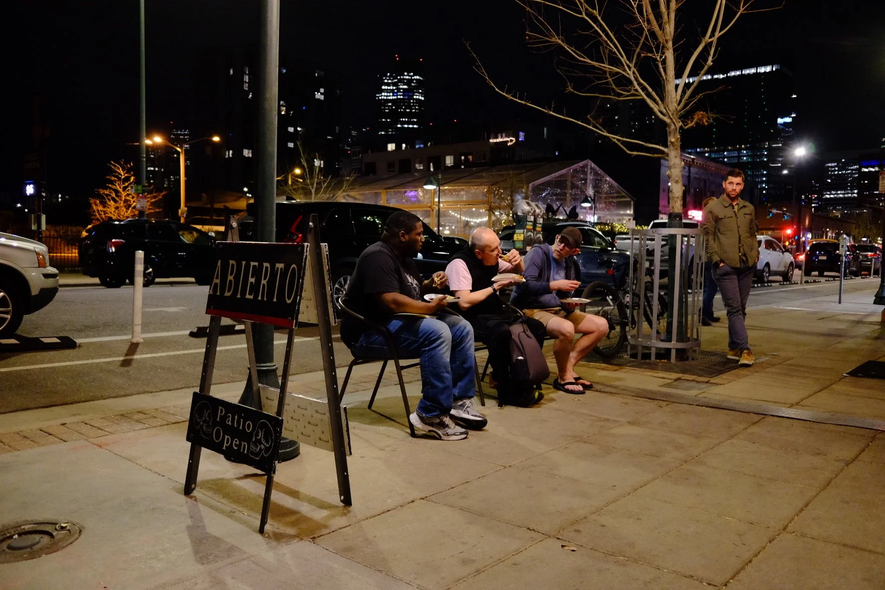 Night scene of city street with three men sitting on chairs eating, one man standing near a tree, parked cars, and city lights in the background.