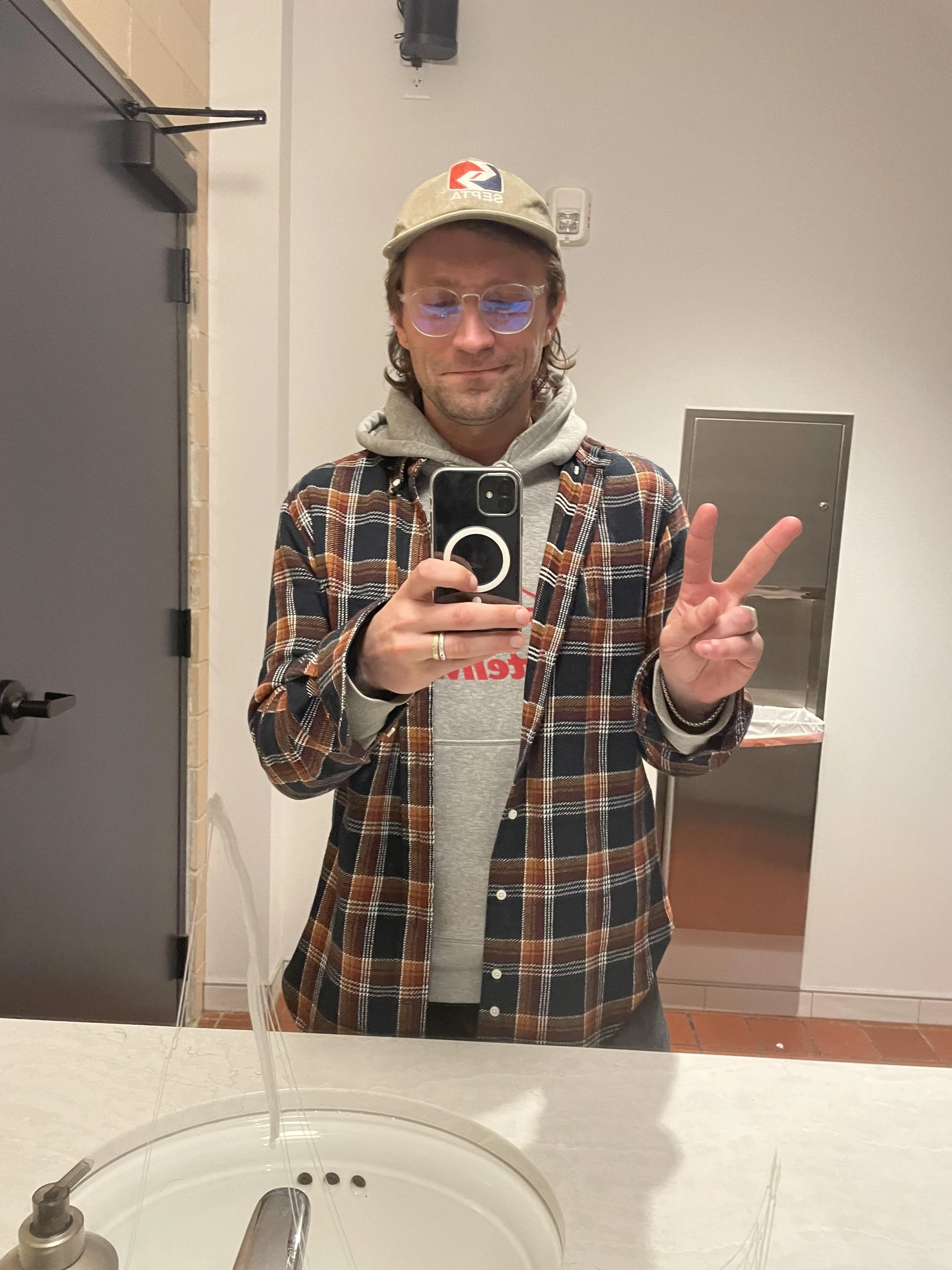 A man in a beige cap, glasses, gray hoodie, and plaid shirt taking a selfie in a bathroom mirror, making a peace sign with his right hand.