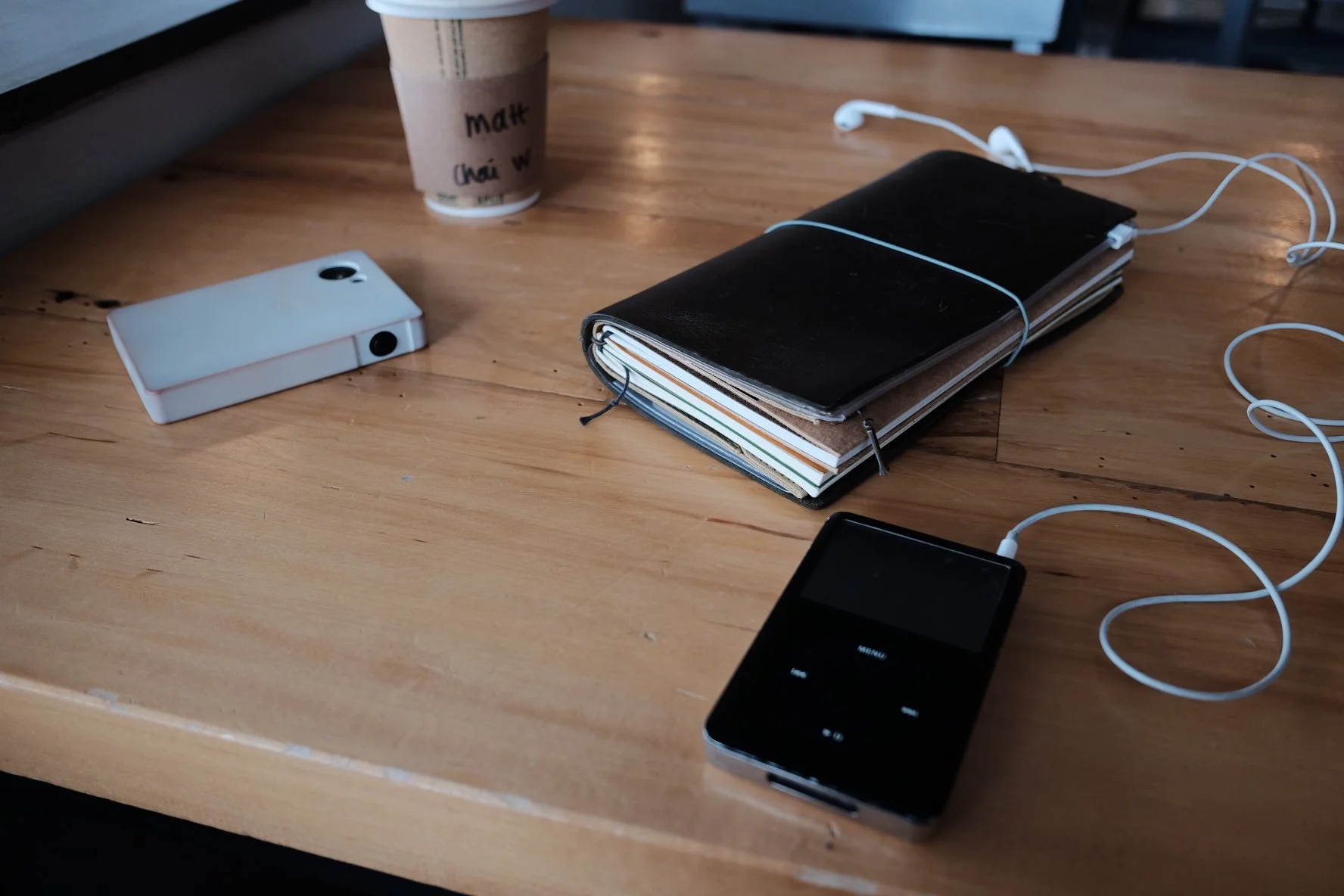 A wooden table with a white dumbphone lying face down, an iPod classic, a travelers notebook bound with a rubber band, a pair of white earphones, and a partially visible paper coffee cup.