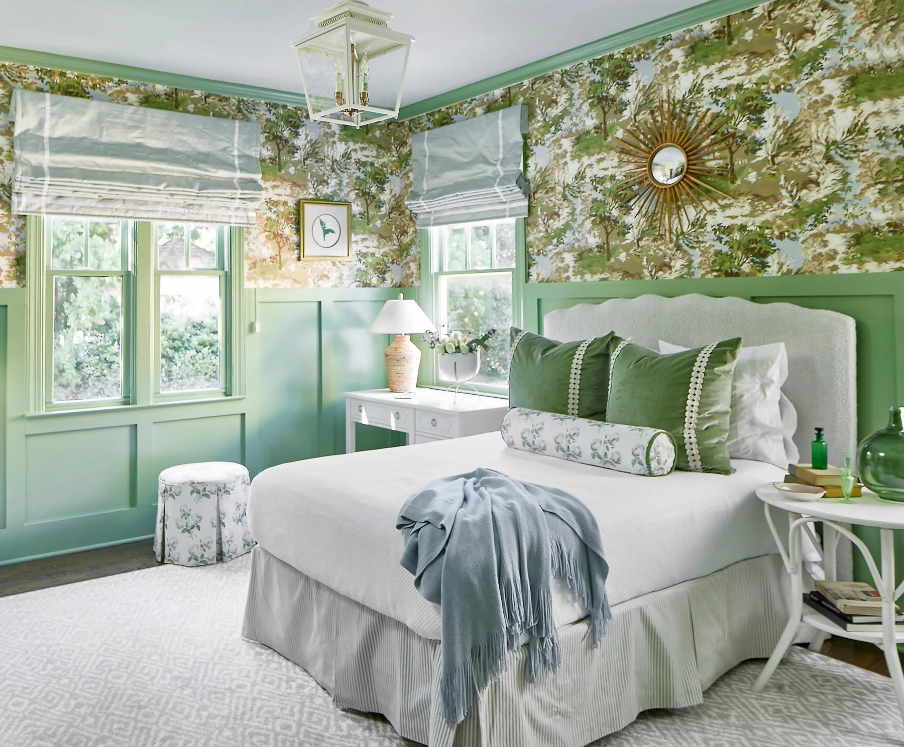 A bedroom with green walls and floral wallpaper, featuring a white bed with green and floral pillows, a white side table with decorative items, floral curtains, and a window with green trim. There is a unique starburst mirror on the wall and a white vanity with a table lamp and flowers, creating a cozy, vintage-inspired decor.