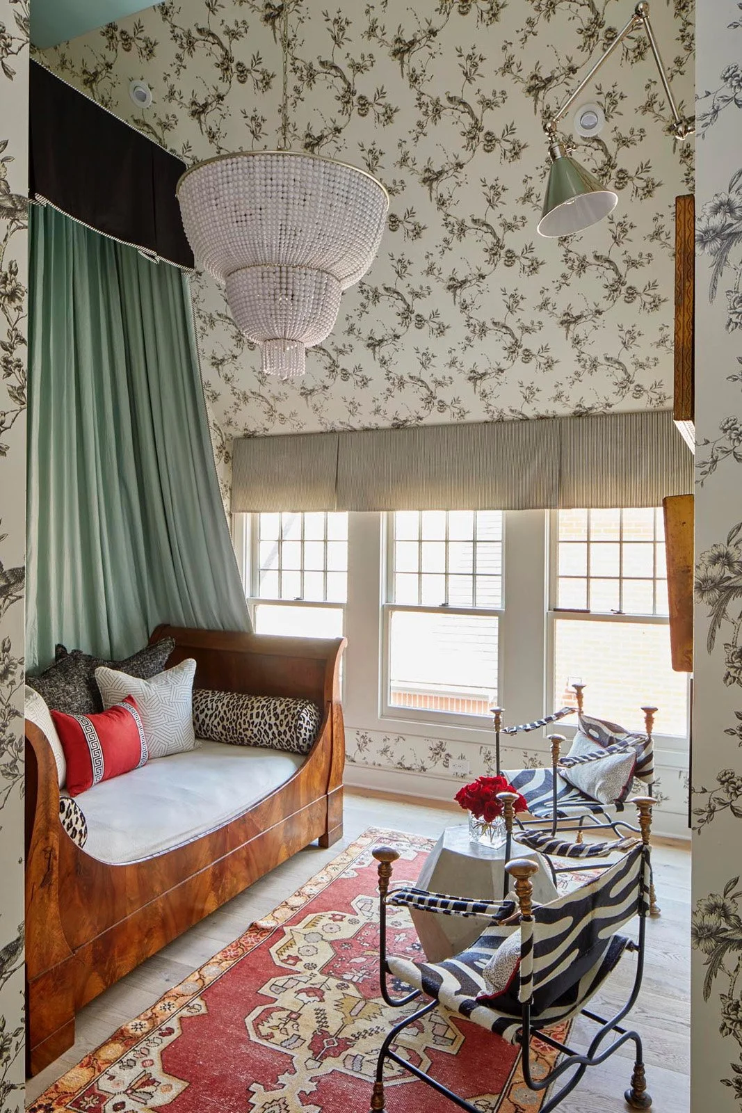 Interior of a cozy living room with floral wallpaper, a large window with blinds, a vintage wooden settee with patterned pillows, with an energetic zigzag patterned chair and a floral vase on a small table, a colorful rug, and a chandelier hanging from the ceiling.