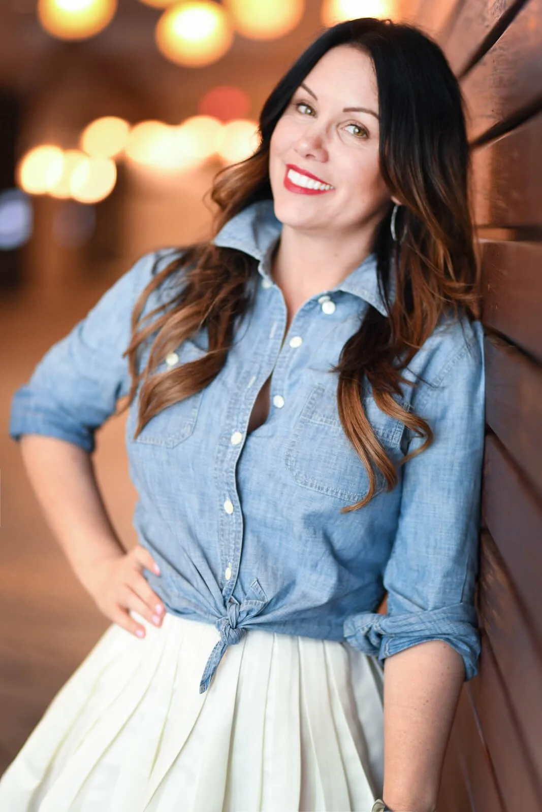 A woman with dark hair and green eyes smiling, leaning against a wooden wall, wearing a blue button-up shirt tied at the waist and a white pleated skirt, with blurred warm lights in the background.