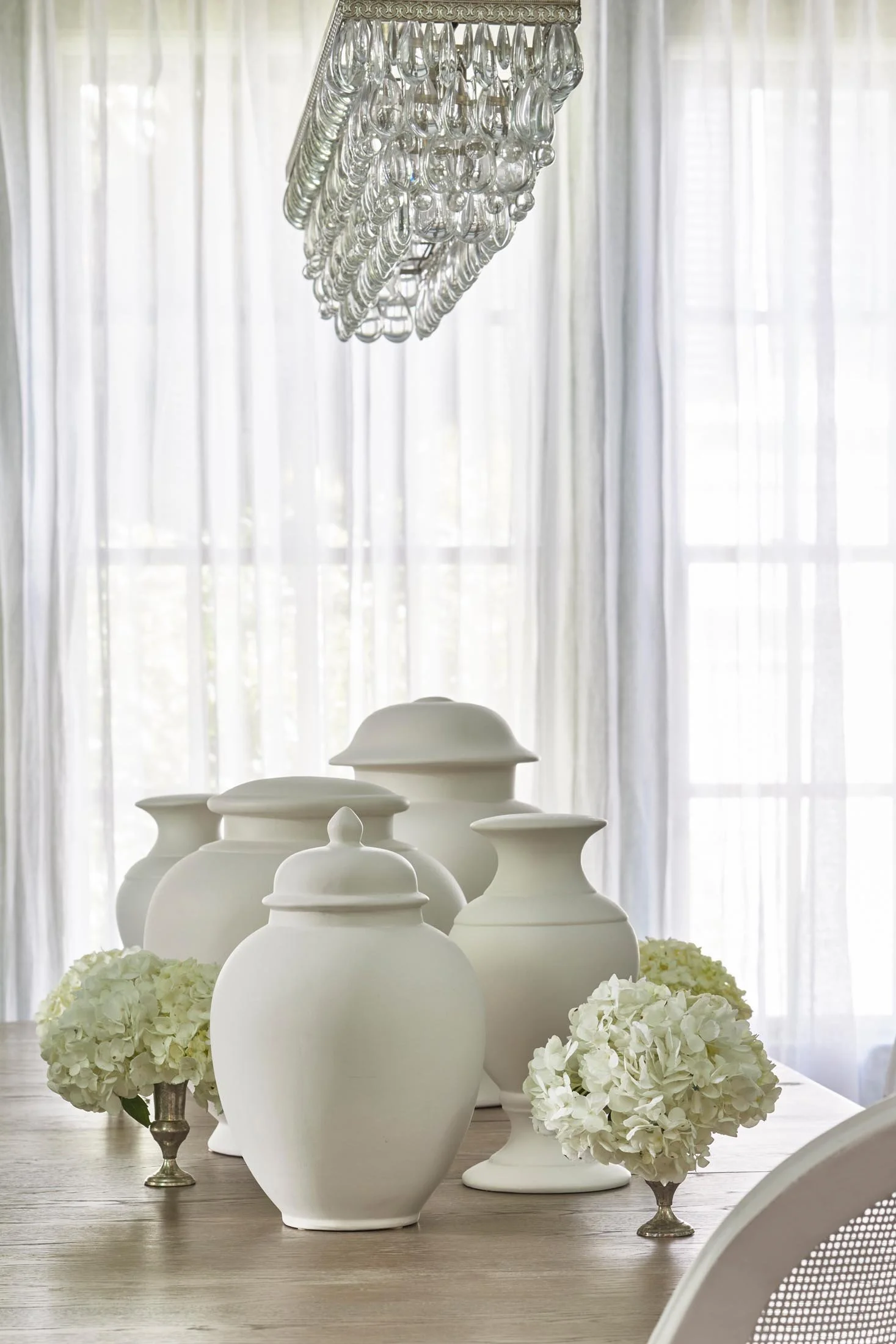 A dining table decorated with white ceramic vases and white hydrangea flowers, set in front of sheer white curtains with a crystal chandelier overhead.