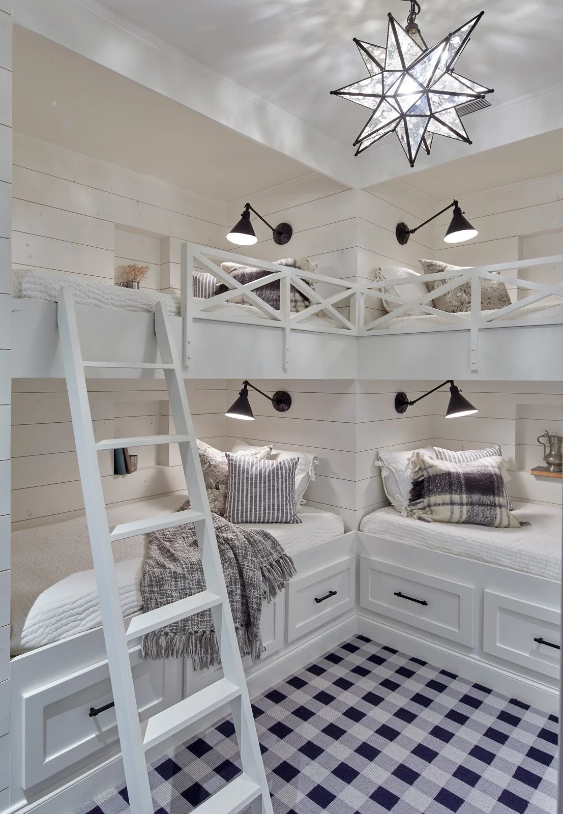 A cozy attic bedroom with white paneled walls, black wall-mounted lamps, and a ceiling star-shaped chandelier. There are two beds with pillows and throws, a white ladder, and a black and white checkered rug.