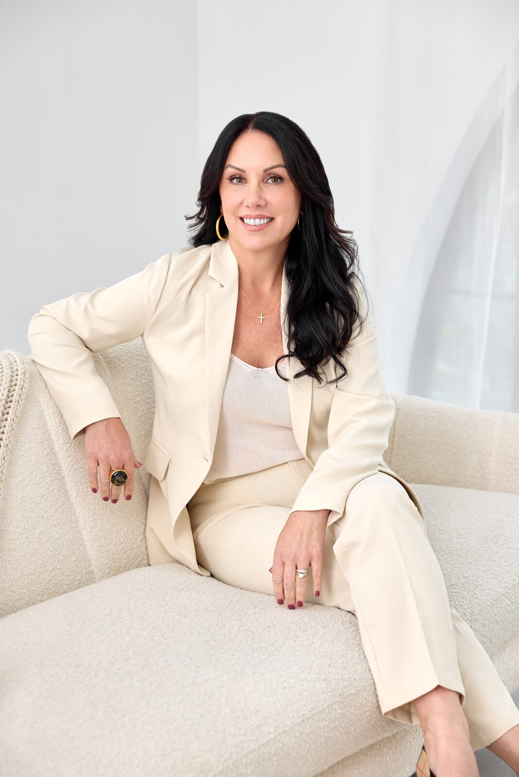 A woman with long black hair, wearing a cream-colored suit and jewelry, sitting on a cream-colored sofa in a bright room and smiling at the camera.