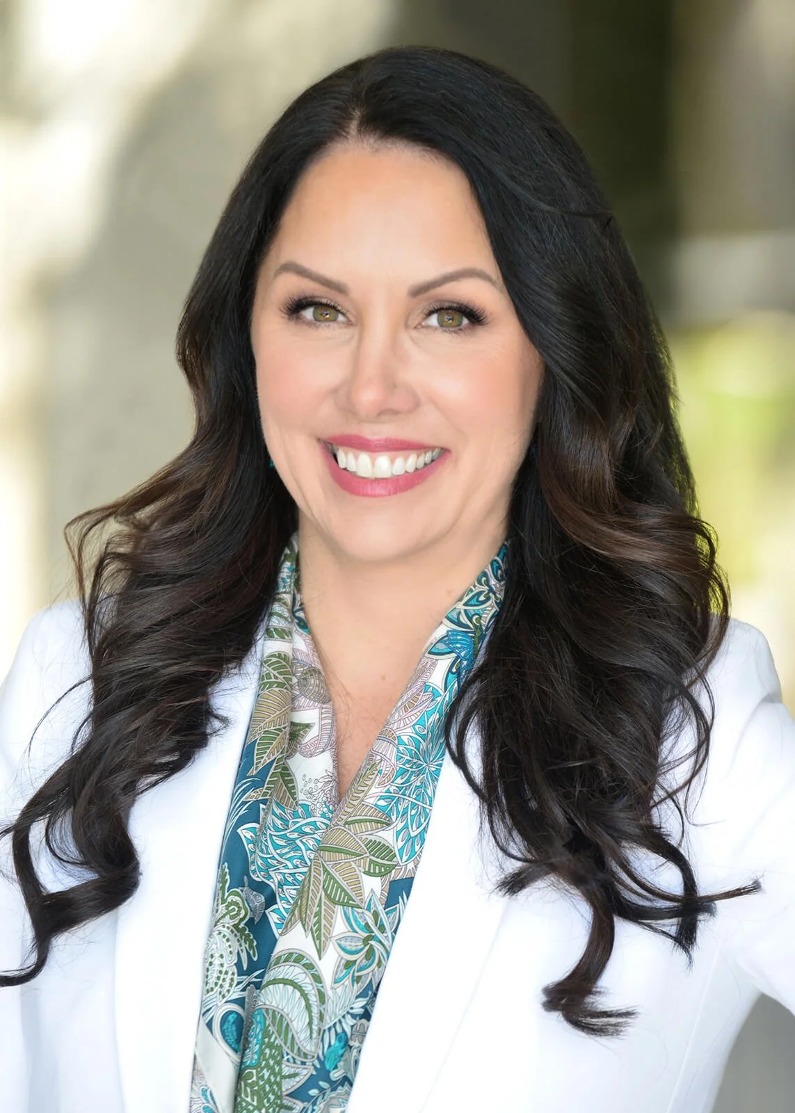 A woman with long, curly dark hair and green eyes smiling, wearing a white blazer and a colorful patterned scarf, standing outdoors with blurred greenery in the background.
